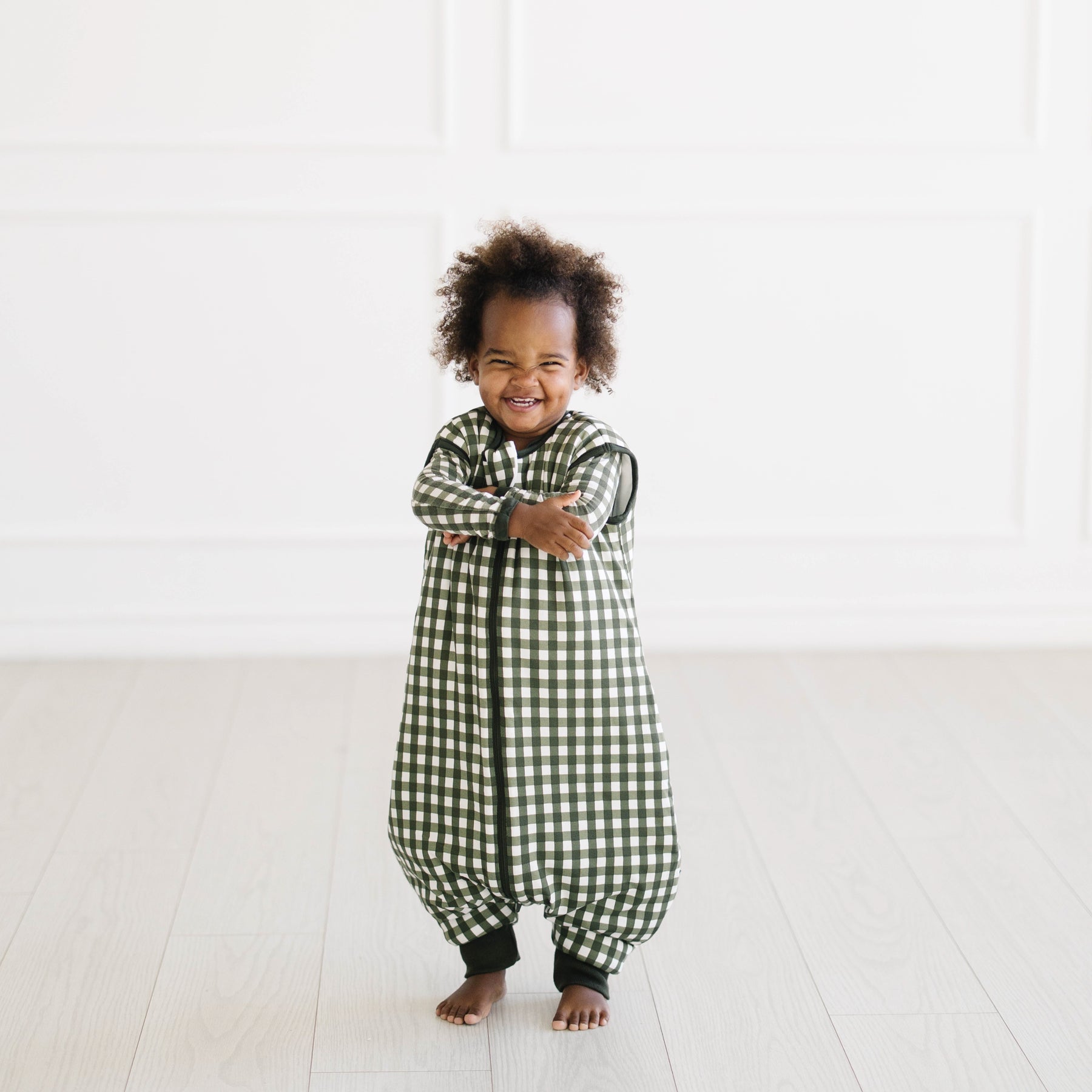 Young toddler standing in front of a white wall wearing the Sleep Bag Walker in Gingham Fir 2.5