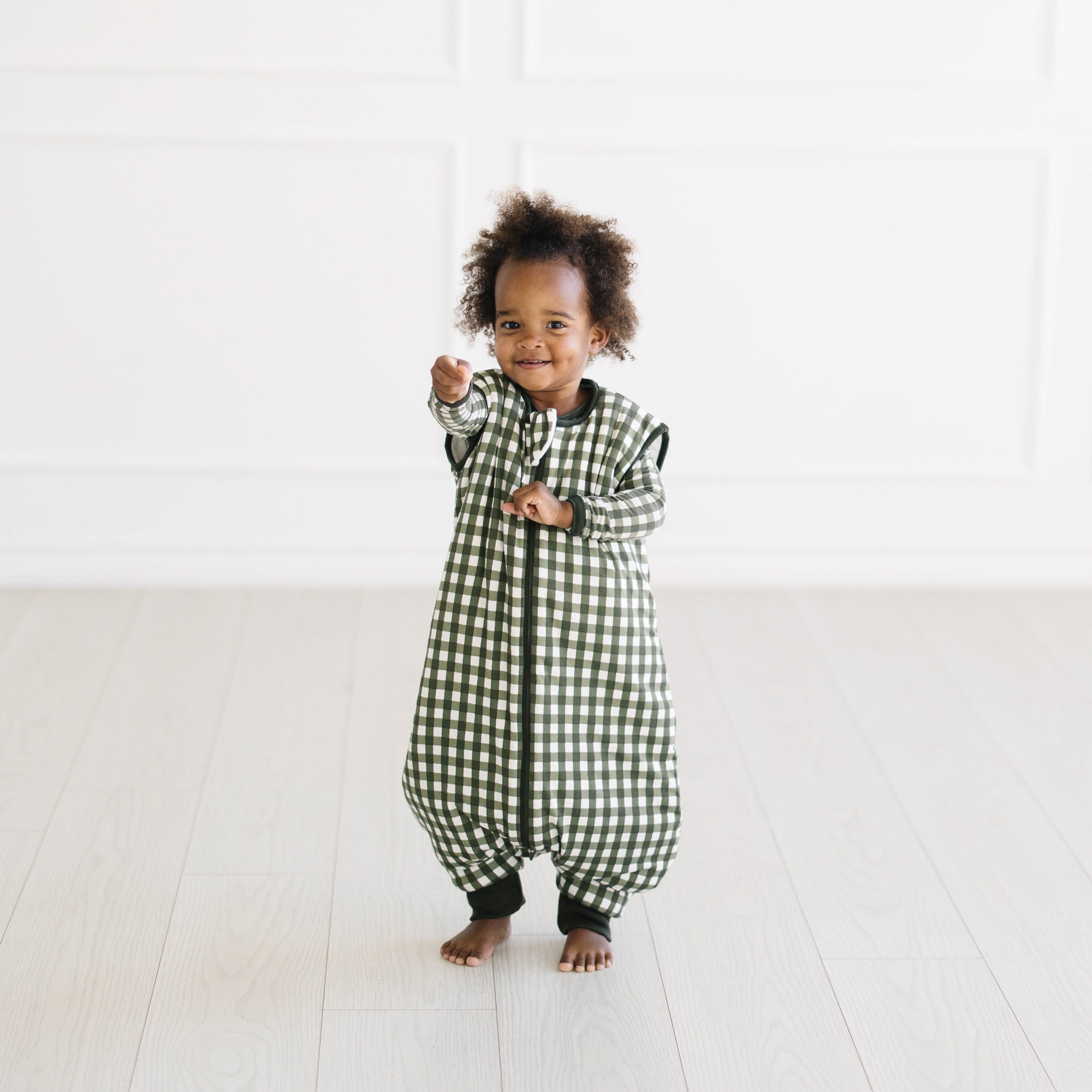 Young toddler standing wearing the Sleep Bag Walker in Gingham Fir 2.5 with matching long sleeve toddler pajamas on underneath