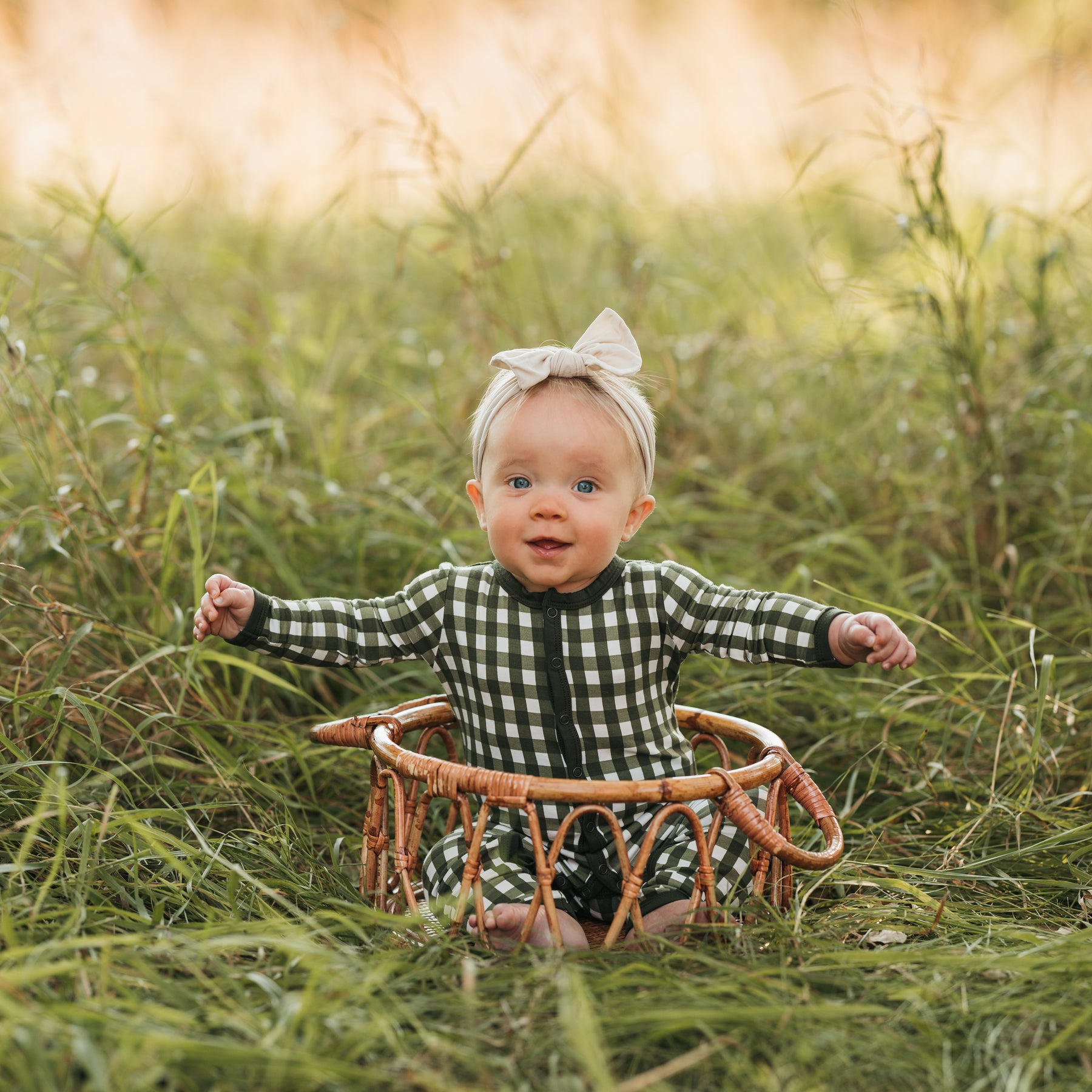 Young infant girl sitting in a wicker basket in a field wearing the Romper in Gingham Fir