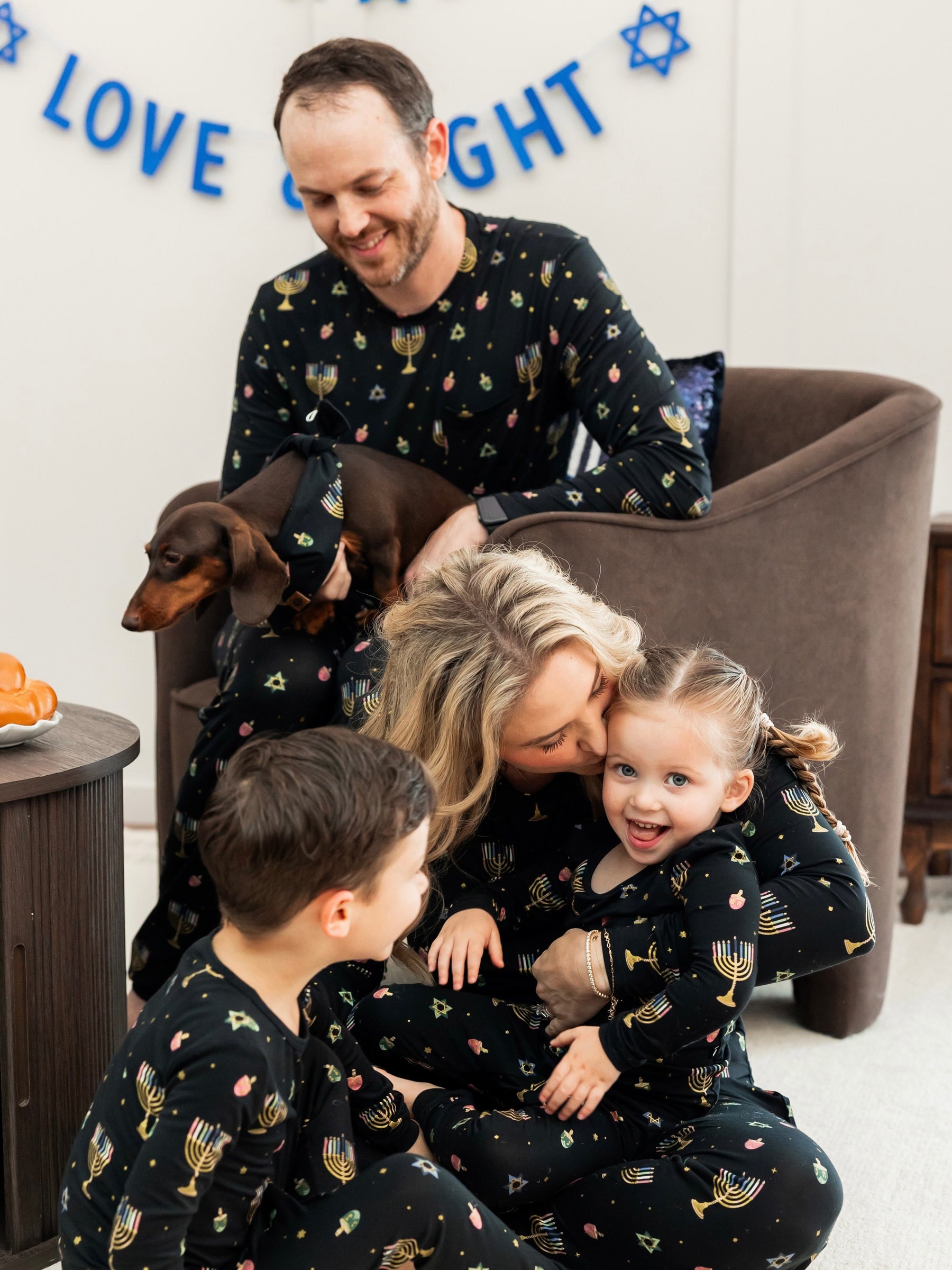 A family of four wearing matching black pajamas with a colorful menorah and Star of David pattern, gathered in a cozy living room setting with a brown armchair and a dachshund.