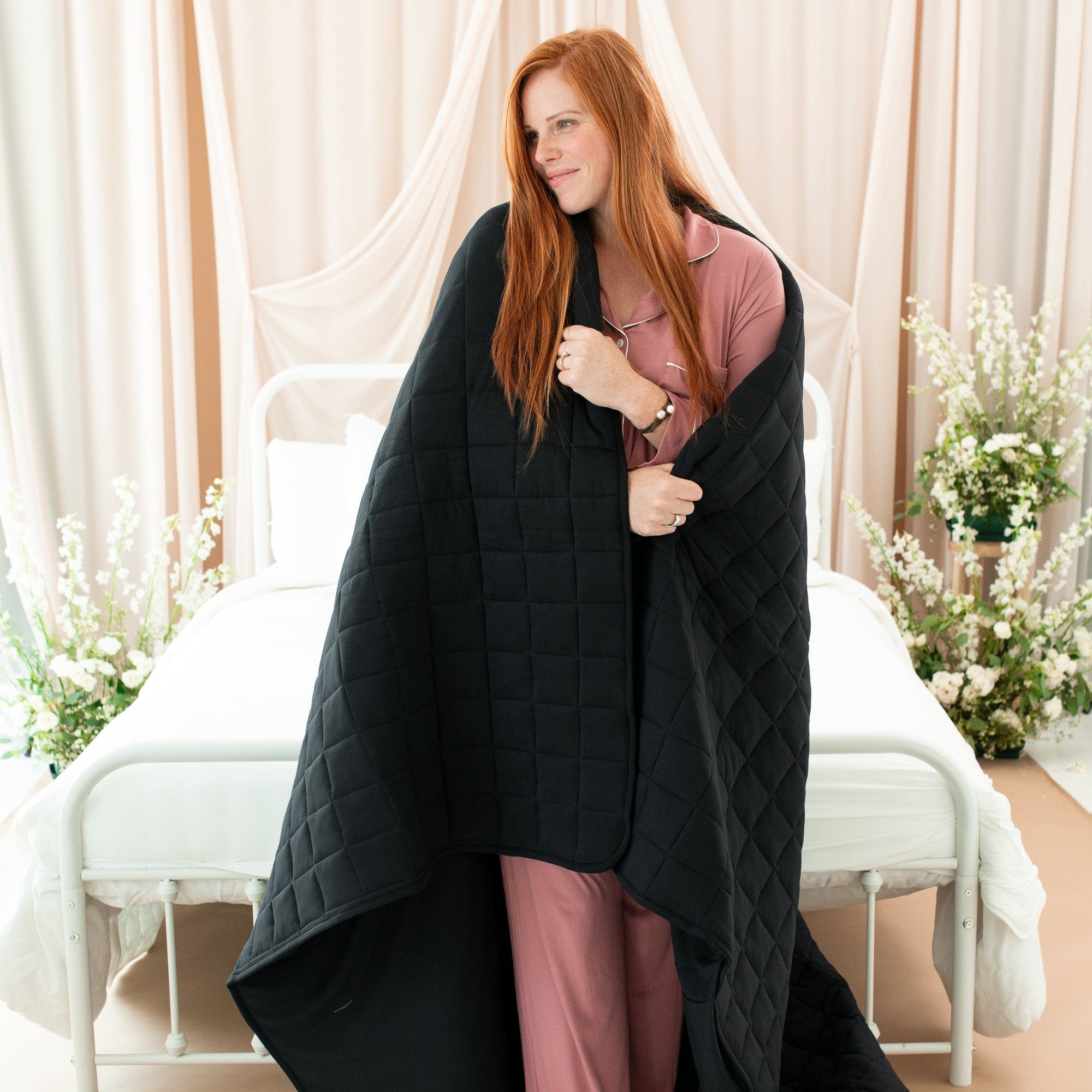 Model wearing dusty rose women's long sleeve pajama set wrapped in Kyte Baby Adult Quilted Blanket in Midnight 3.5