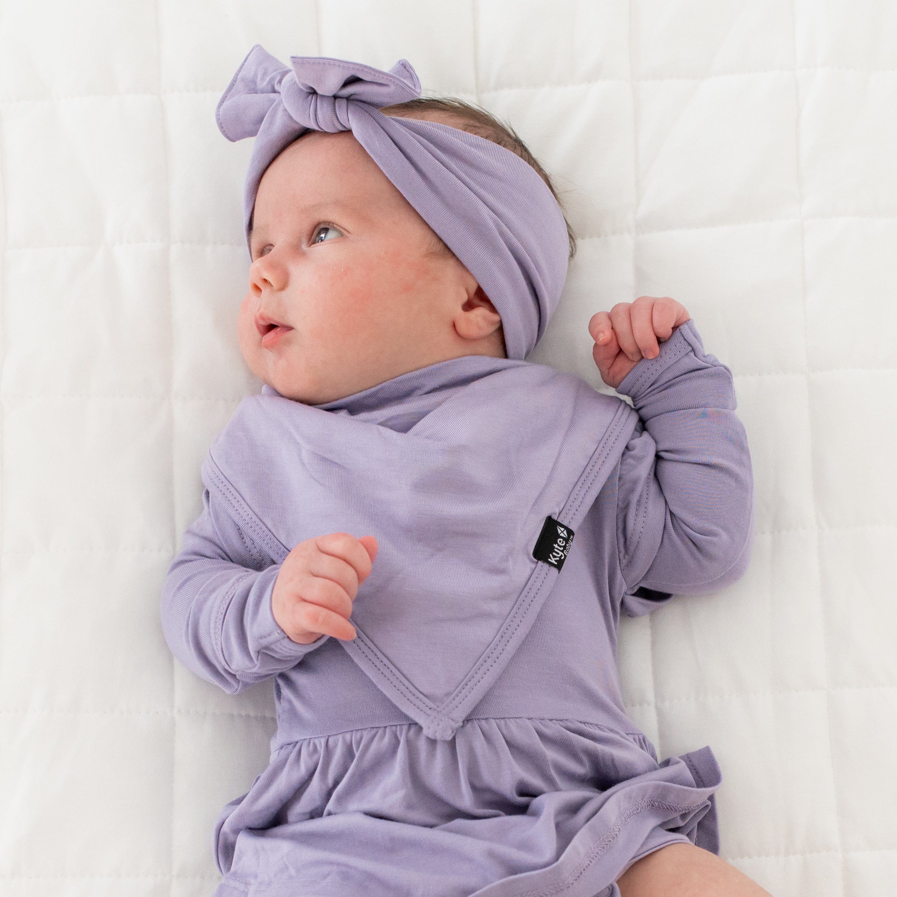 Baby laying on back and wearing Kyte Baby Bow and Matching Bib in Taro