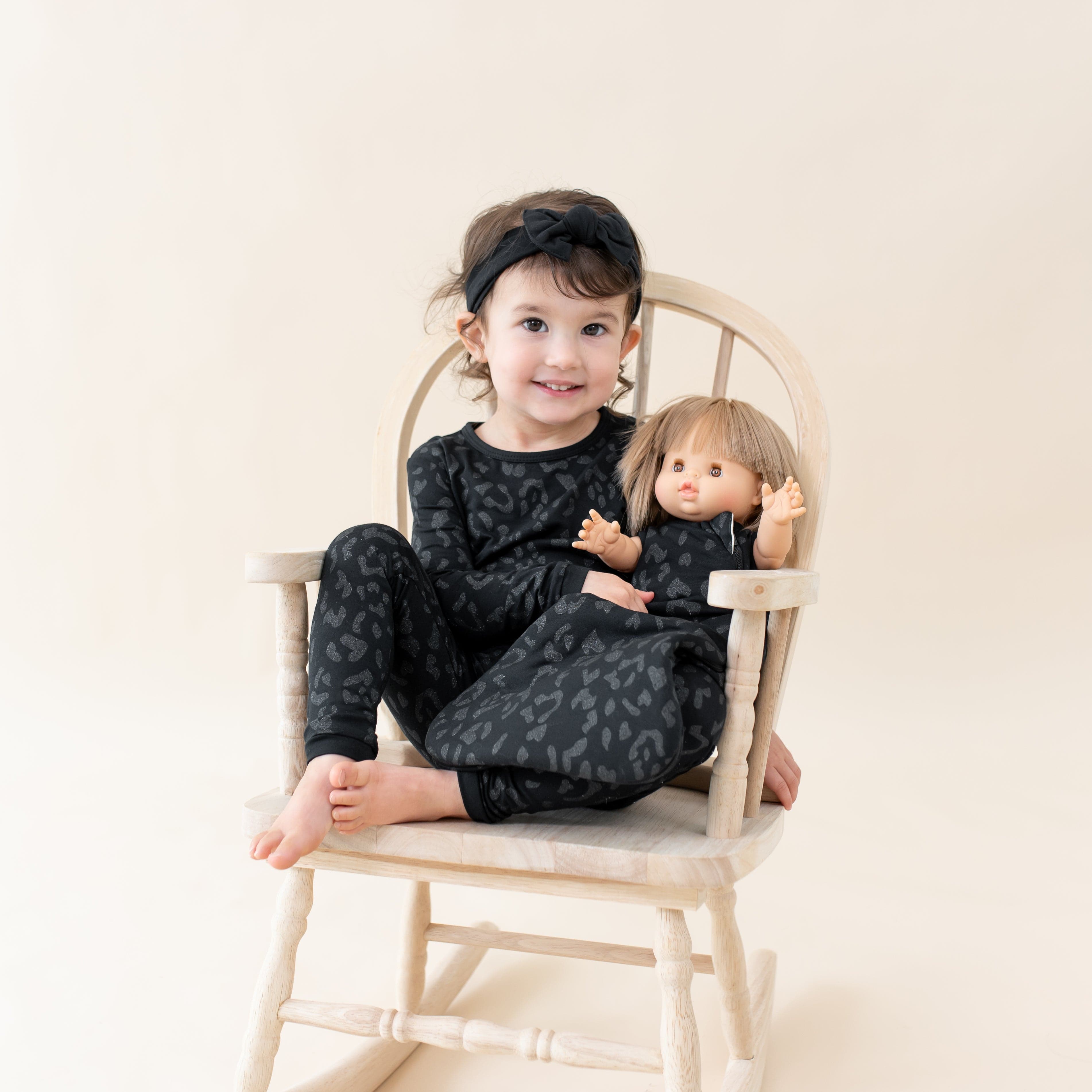 Toddler wearing two piece Kyte Baby Long Sleeve Pajamas in Midnight Leopard with doll in matching sleep sack