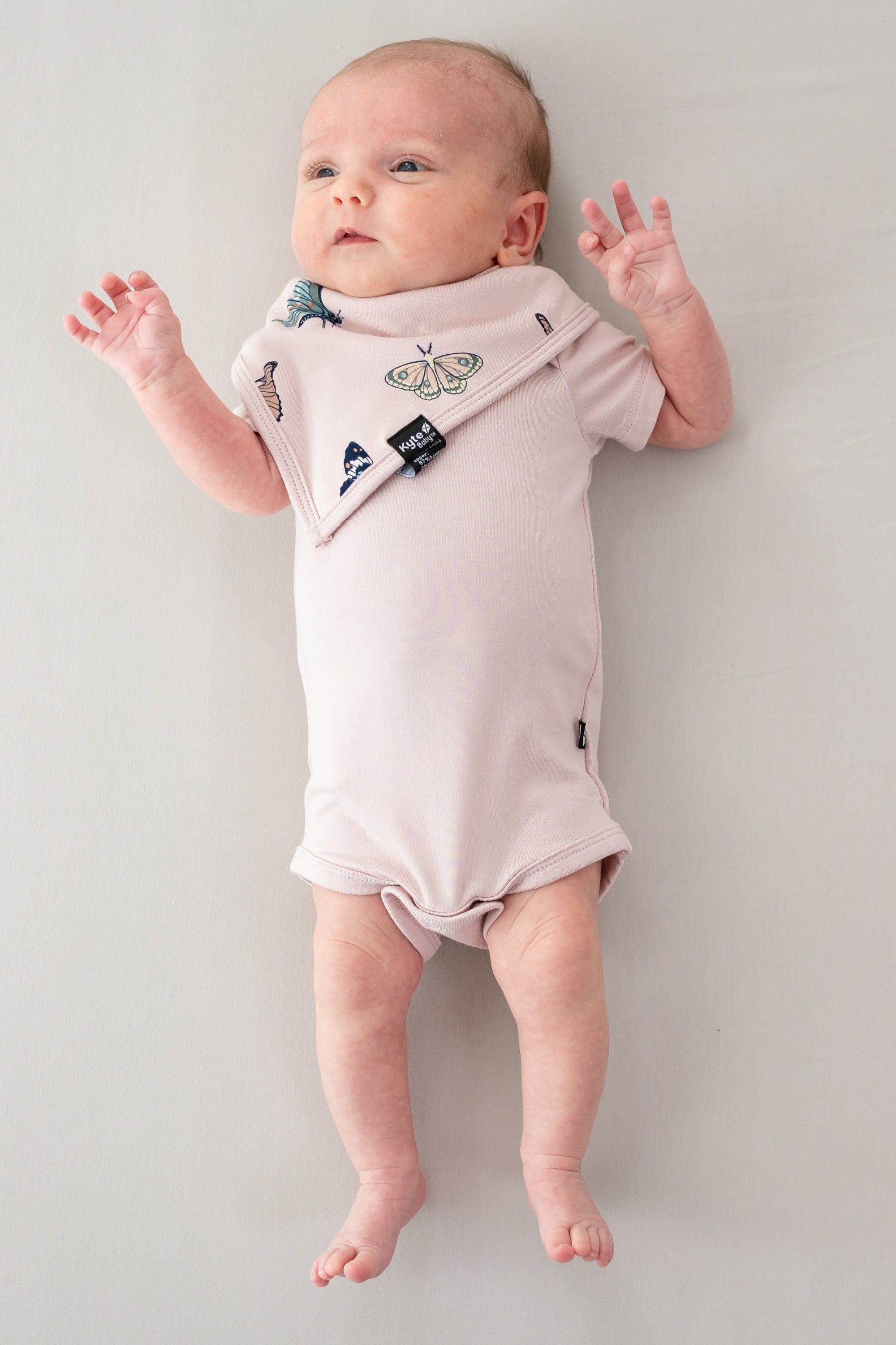 Newborn laying down with hands in the air wearing the Bib in Blush Butterfly and Blush short sleeve bodysuit
