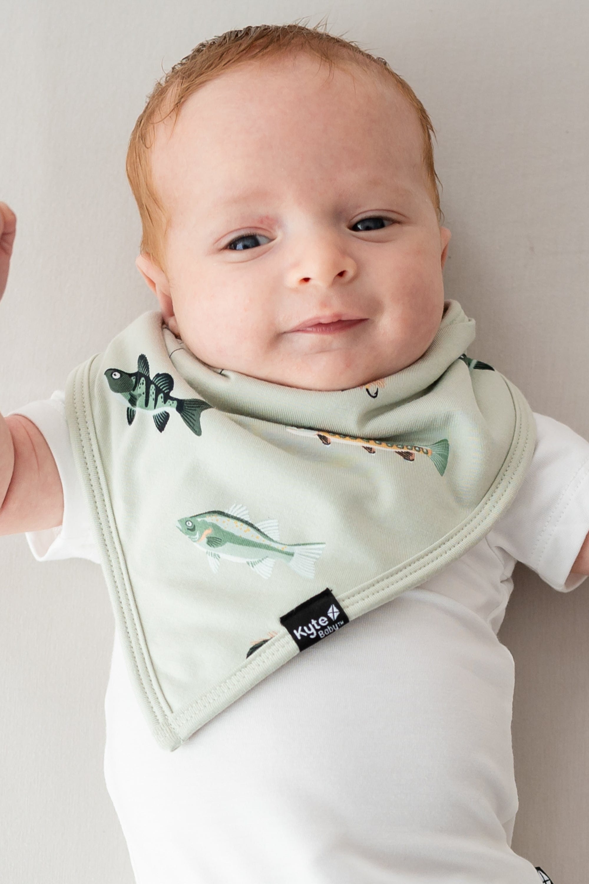 Infant laying down on a light neutral surface wearing the Bib in Fishing and a white bodysuit