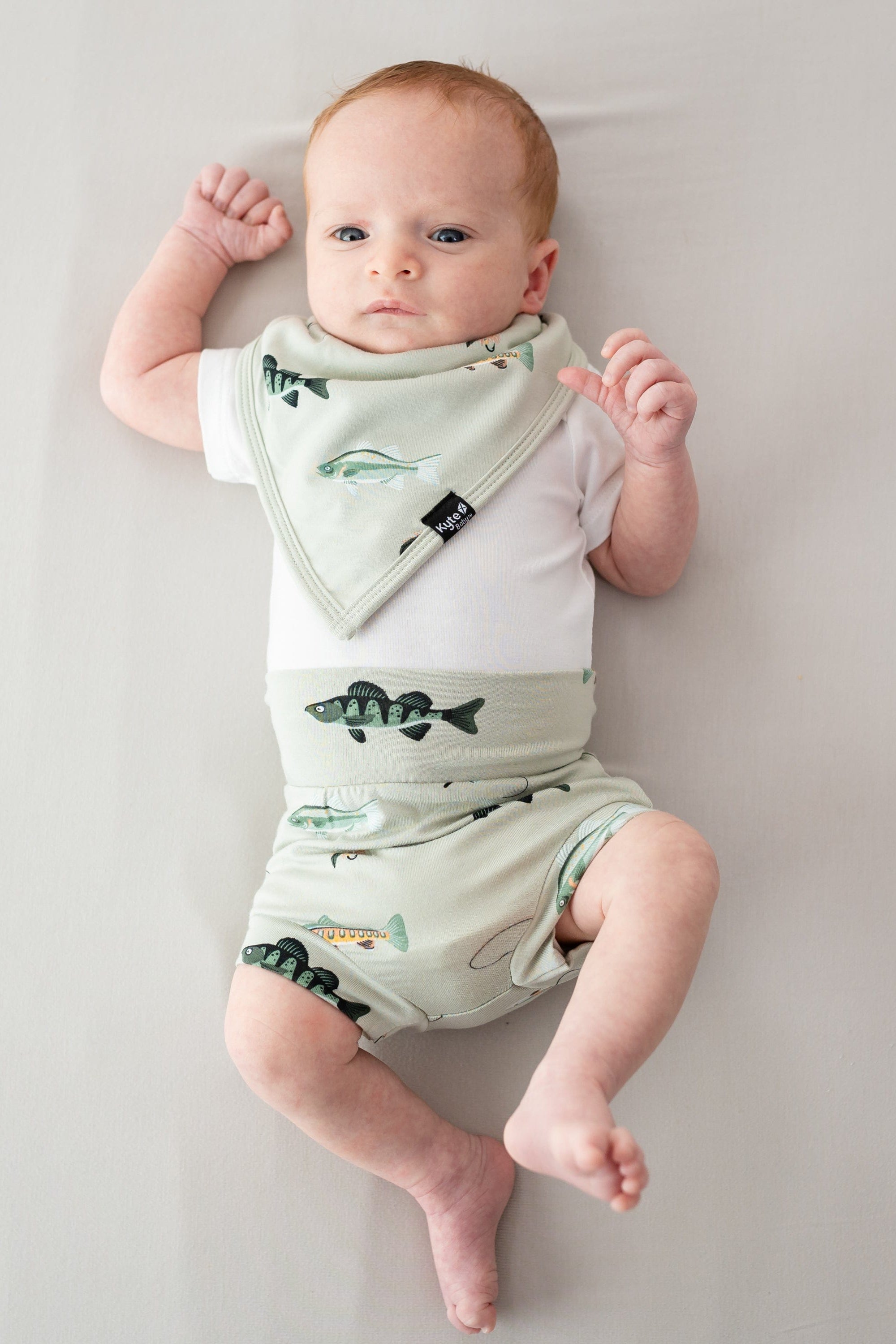Infant laying down on a light neutral surface waring the Bib in Fishing with matching bummies and a white bodysuit