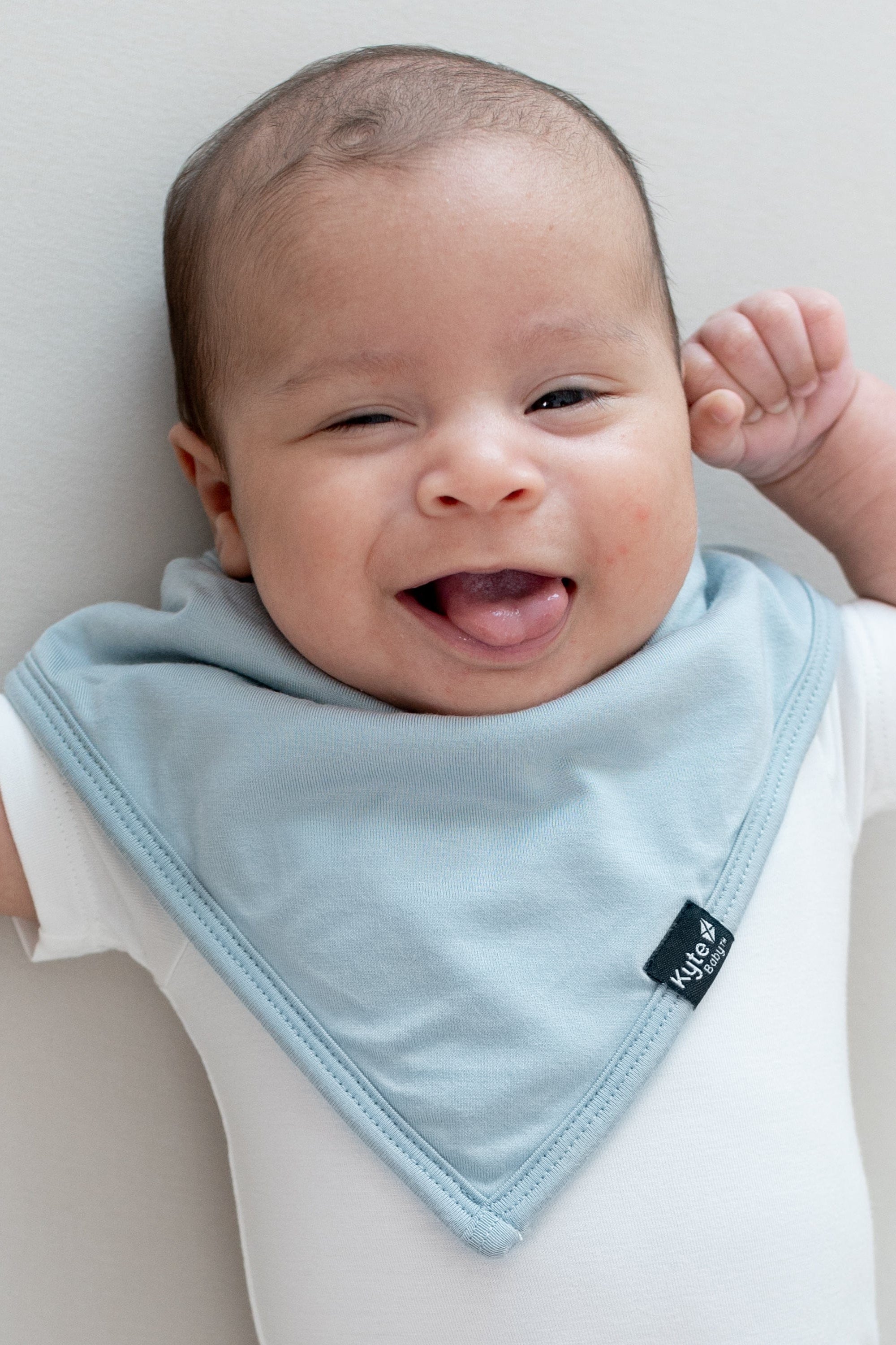 Infant laying down sticking out their tongue wearing the Bib in Fog and a white short sleeve bodysuit
