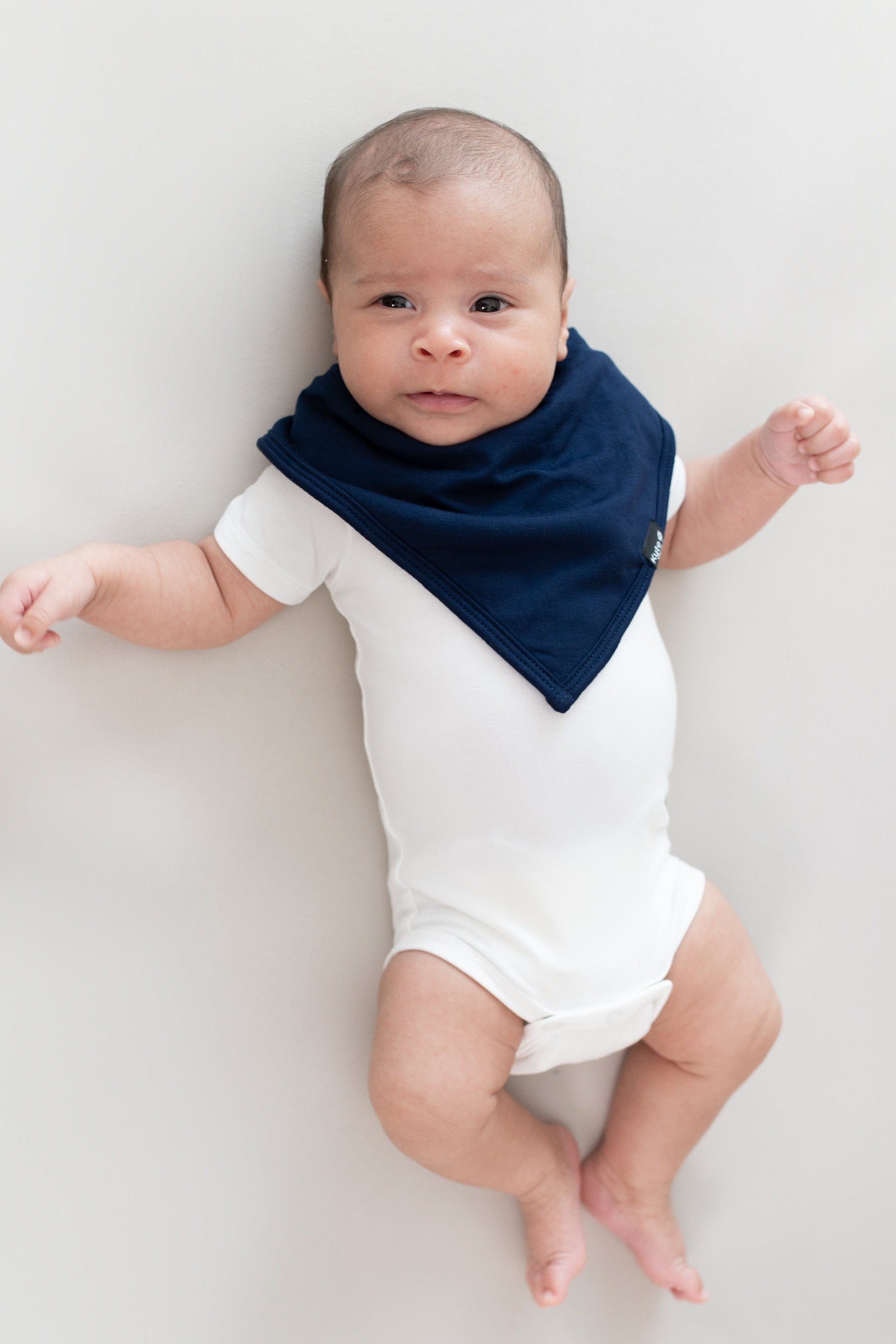 Infant laying on a Crib Sheet in Oat while wearing a Bib in Navy