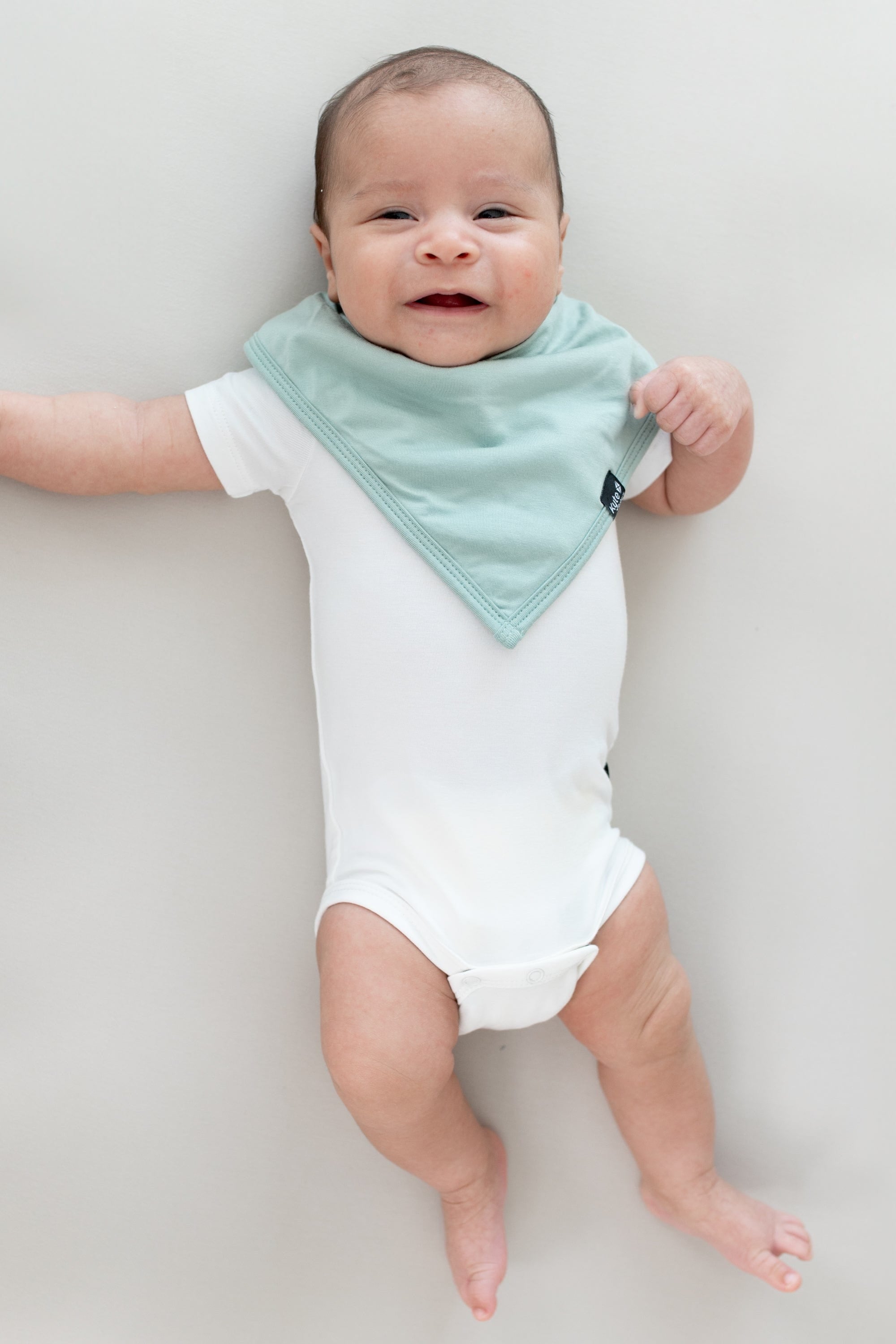 Baby laying on a Crib Sheet in Oat while wearing a  Bib in Sage