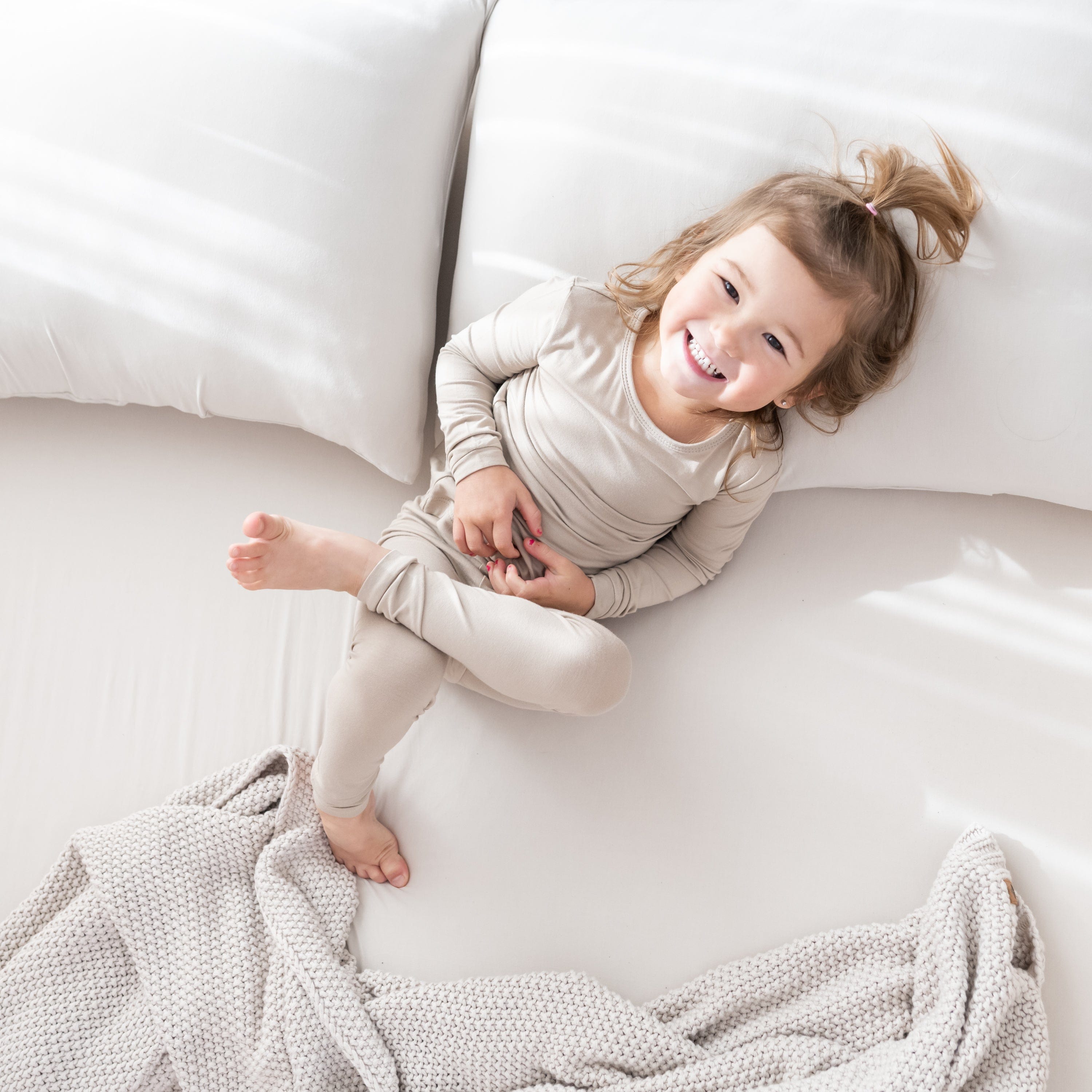 Toddler in Long Sleeve Oat Pajamas laying on a bed with Kyte Baby Fitted Sheet Fitted Sheet with Pillowcases in Almond