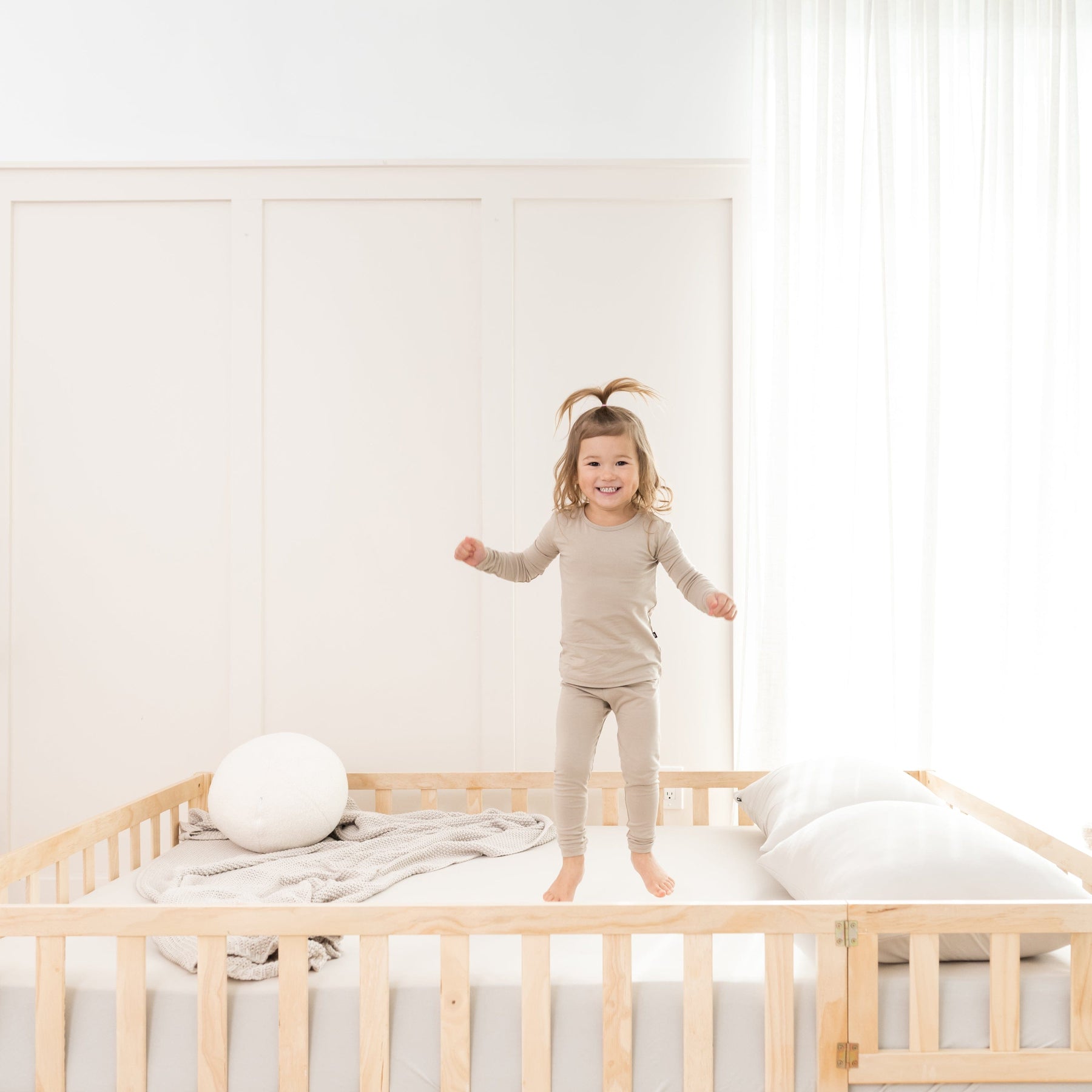 Toddler jumping on bed wearing two piece Kyte Baby Long Sleeve Pajamas in Almond