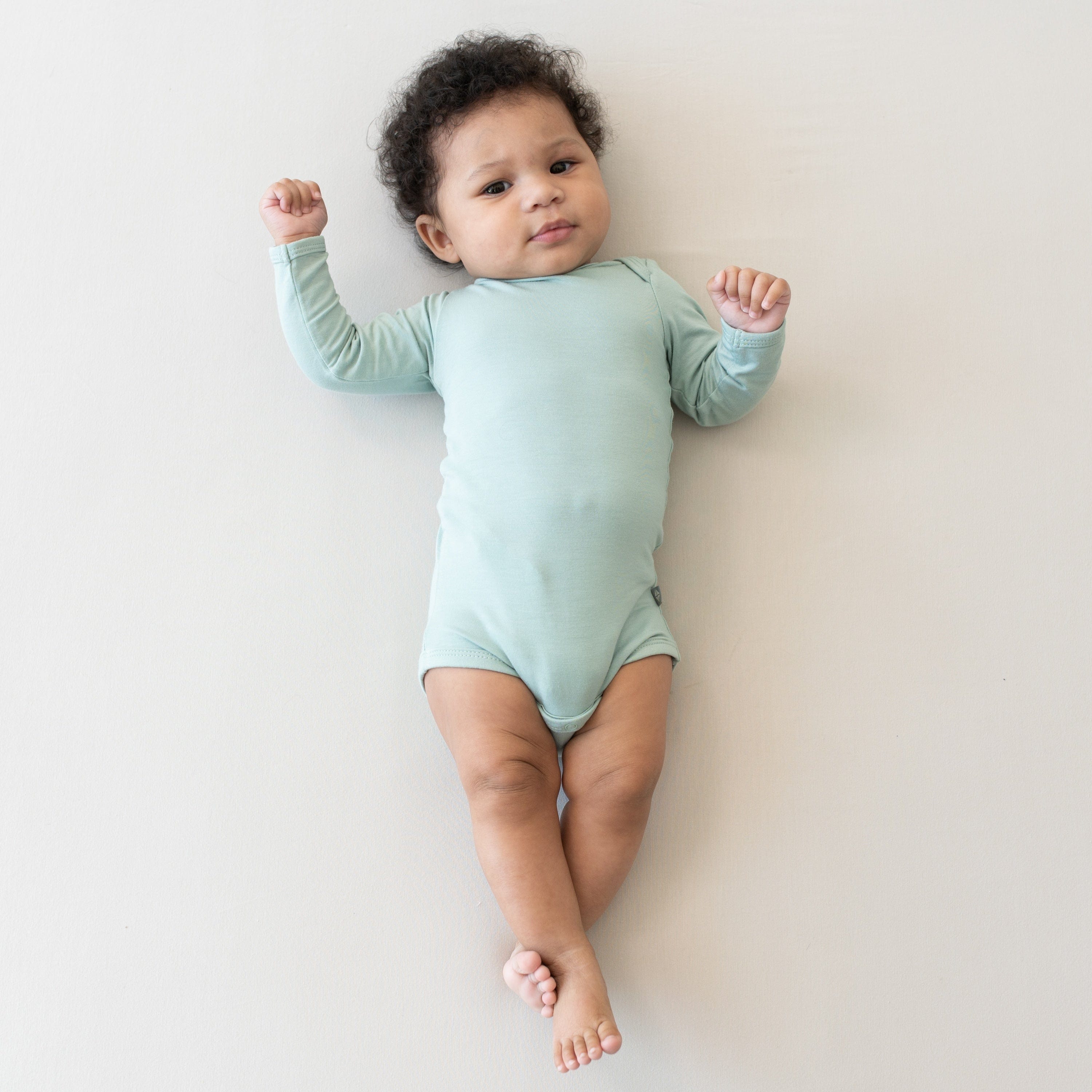 Infant laying on a Crib Sheet in Oat while wearing a Long Sleeve Bodysuit in Sage
