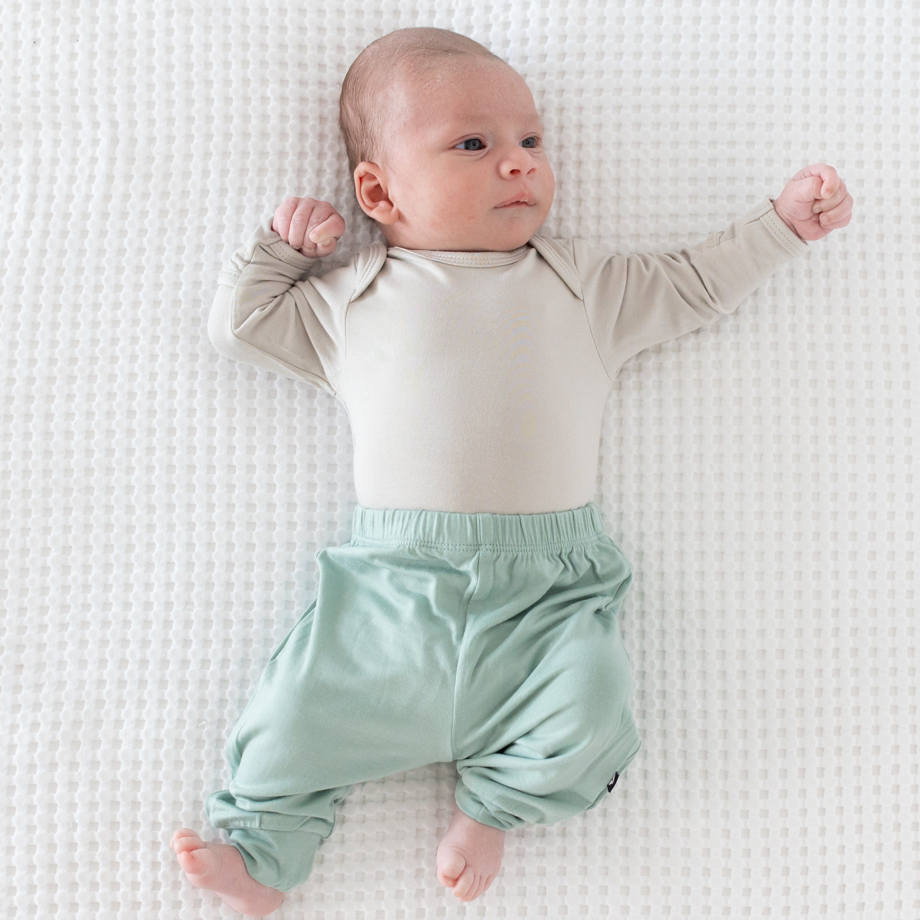 Newborn wearing Kyte Baby Pants in Sage with a Long Sleeve Bodysuit in Oat with one arm extended