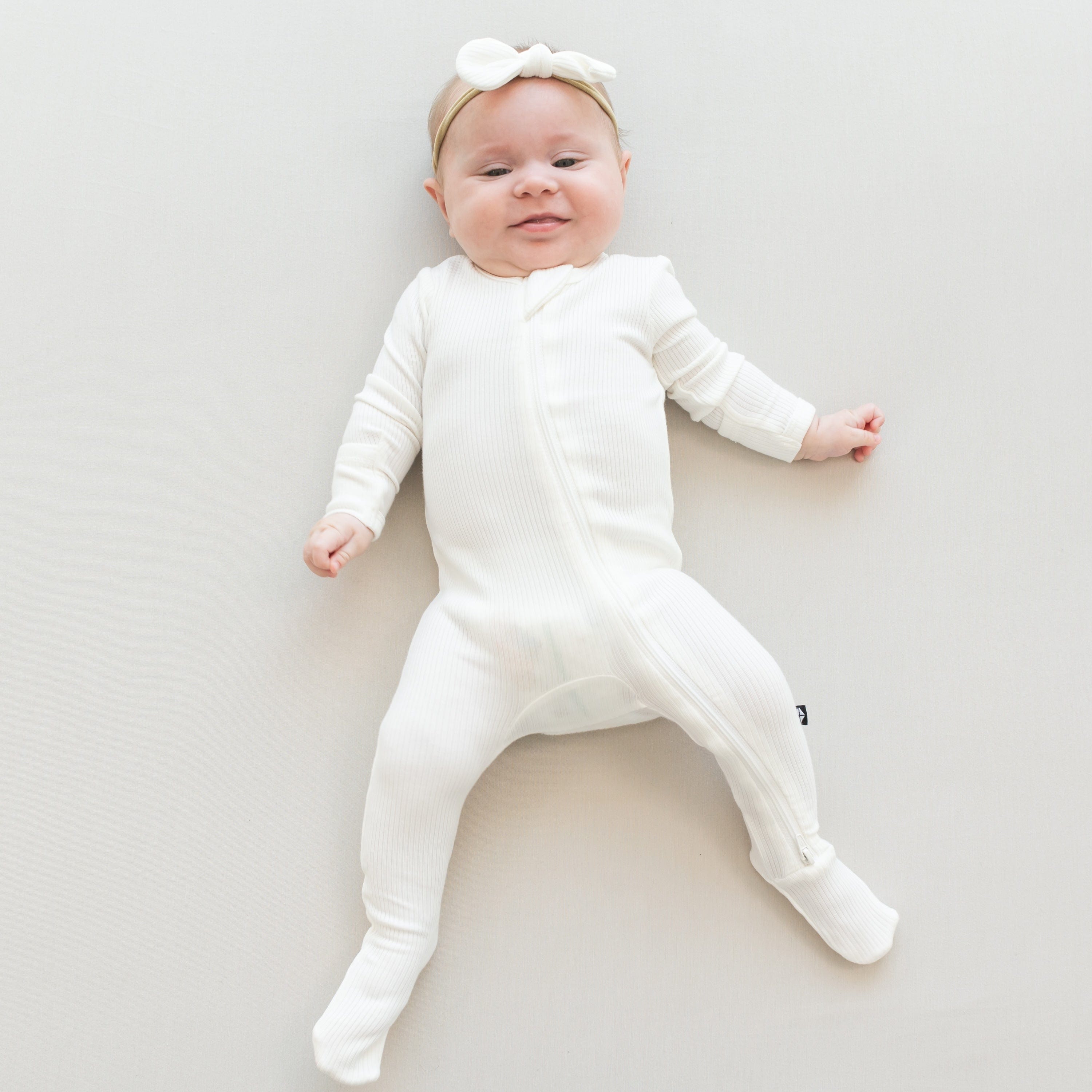 Infant laying on off-white surface wearing Baby Ribbed Zipper Footie Ribbed Zipper Footie in Cloud with a Kyte Baby Bow in Cloud