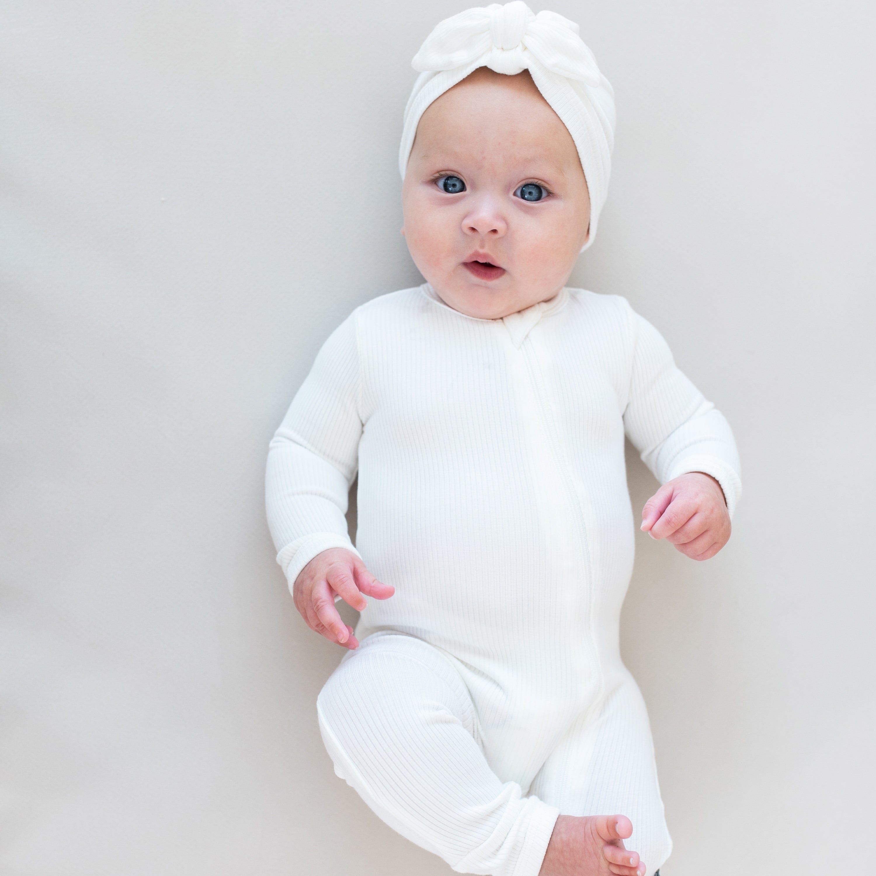 Baby laying on off-white surface wearing Kyte Baby Ribbed Zippered Romper in Cloud and matching Kyte Baby Headwrap