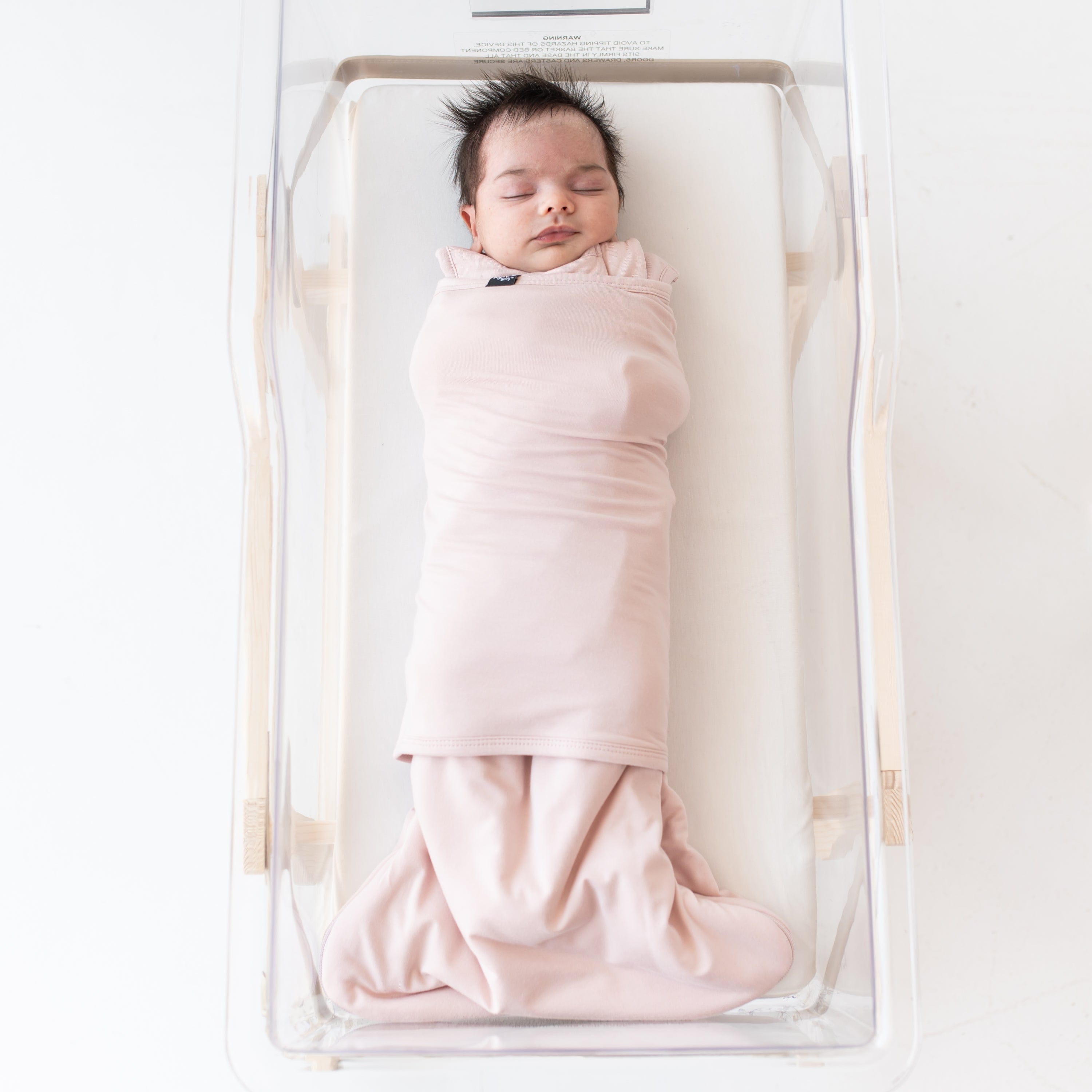 Sleeping baby swaddled in the Sleep Bag Swaddler in Blush 1.0 laying in a bassinet