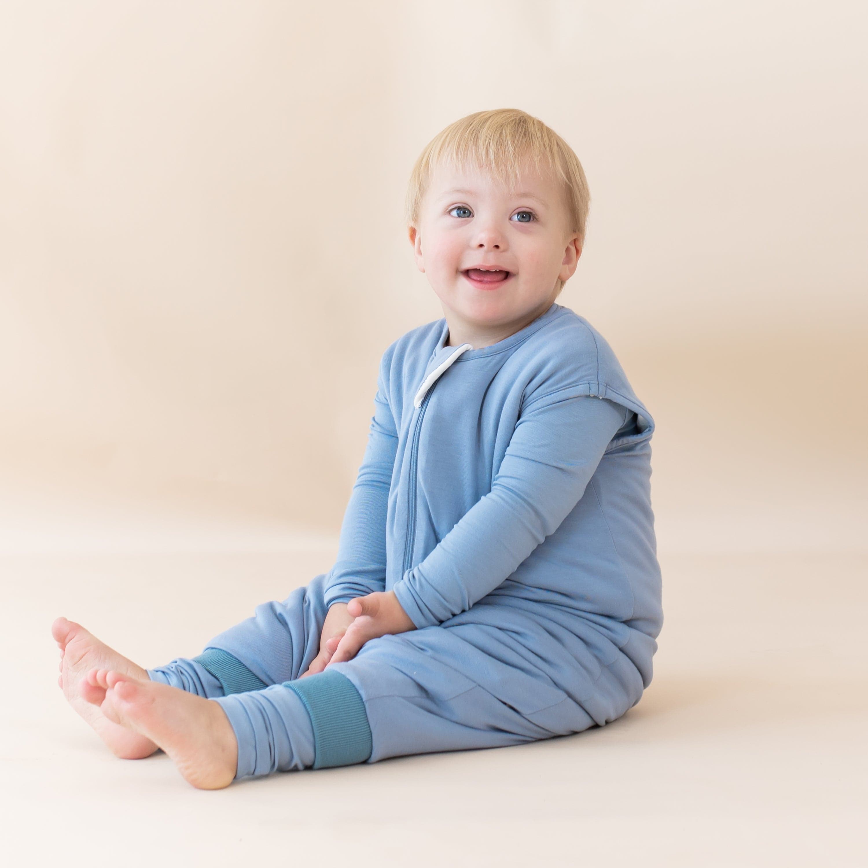 Young child sitting on the floor with legs extended and wearing the Sleep Bag Walker in Slate 1.0
