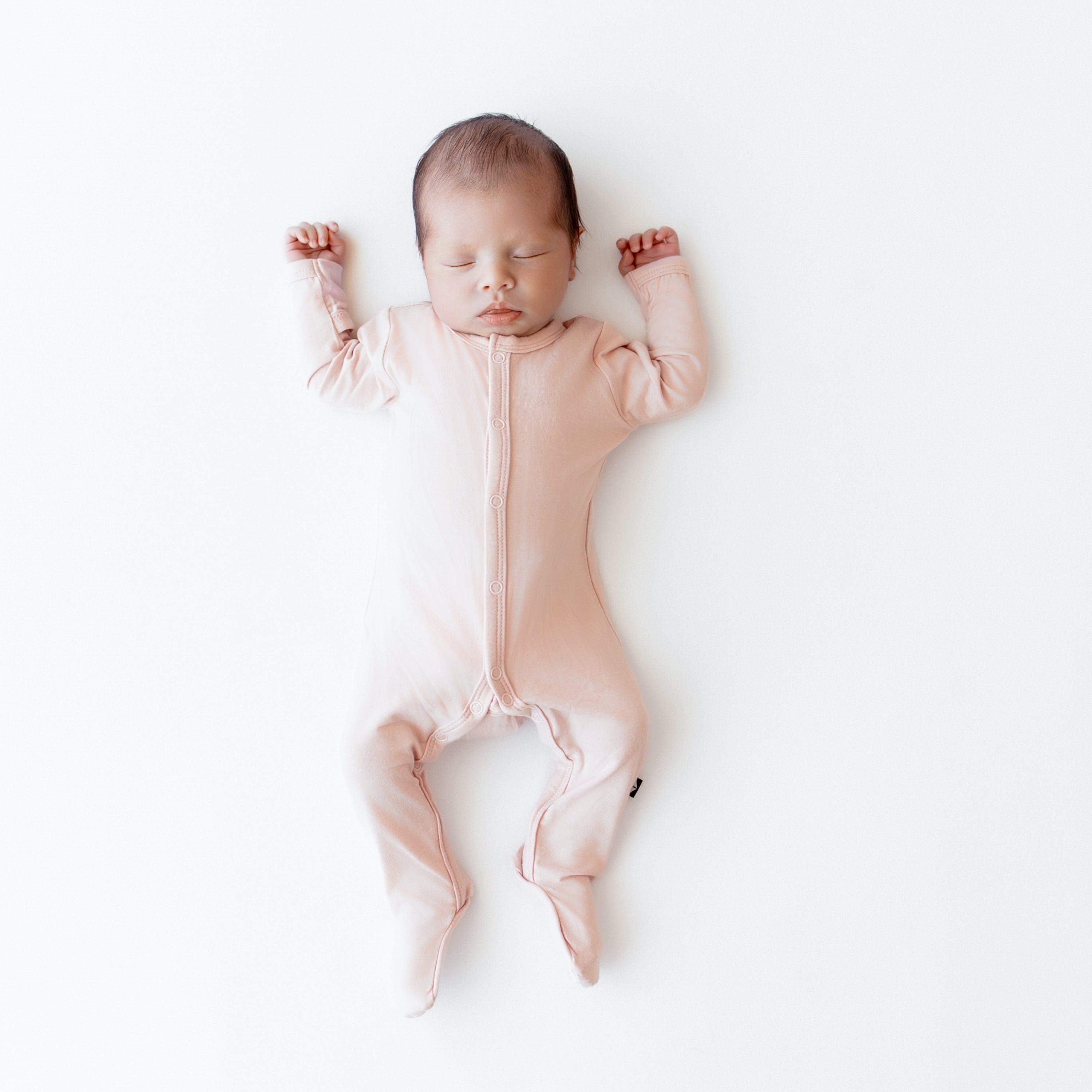 Newborn sleeping on white surface in Kyte Baby Footie in Blush