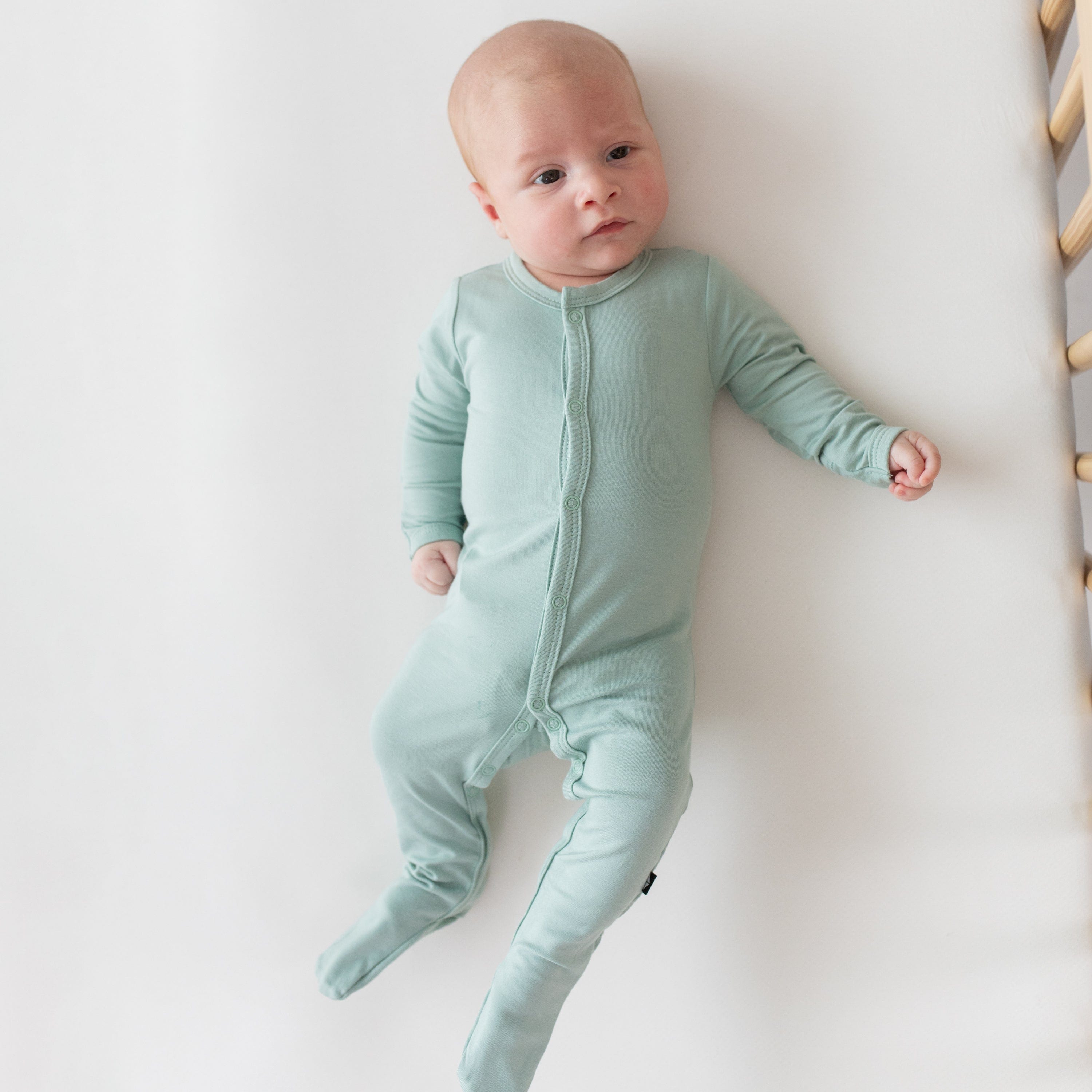 Infant laying on a cribsheet in Oat while wearing Kyte Baby Footie in Sage 