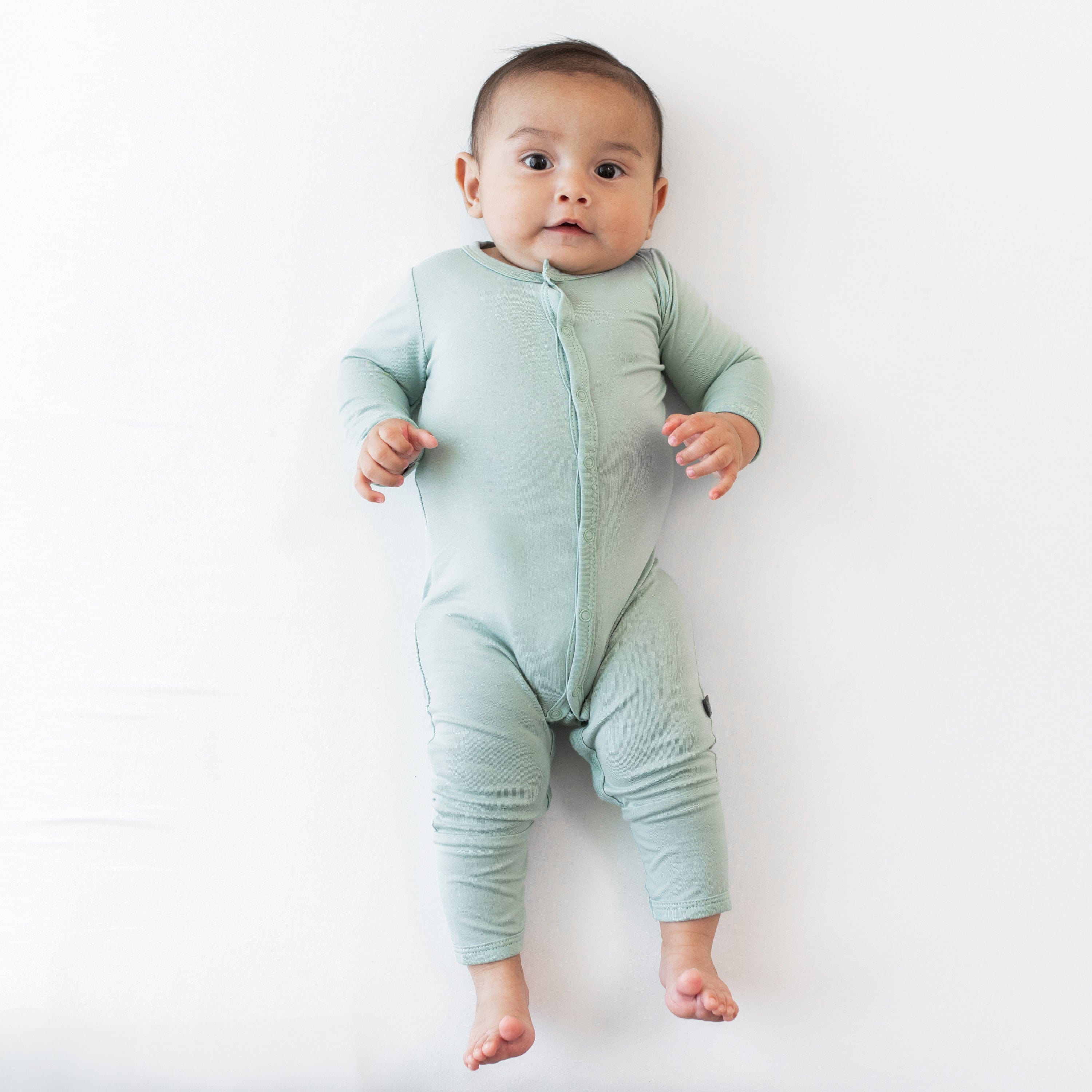 Infant staring at camera while laying on white surface and wearing Kyte Baby Romper in Sage