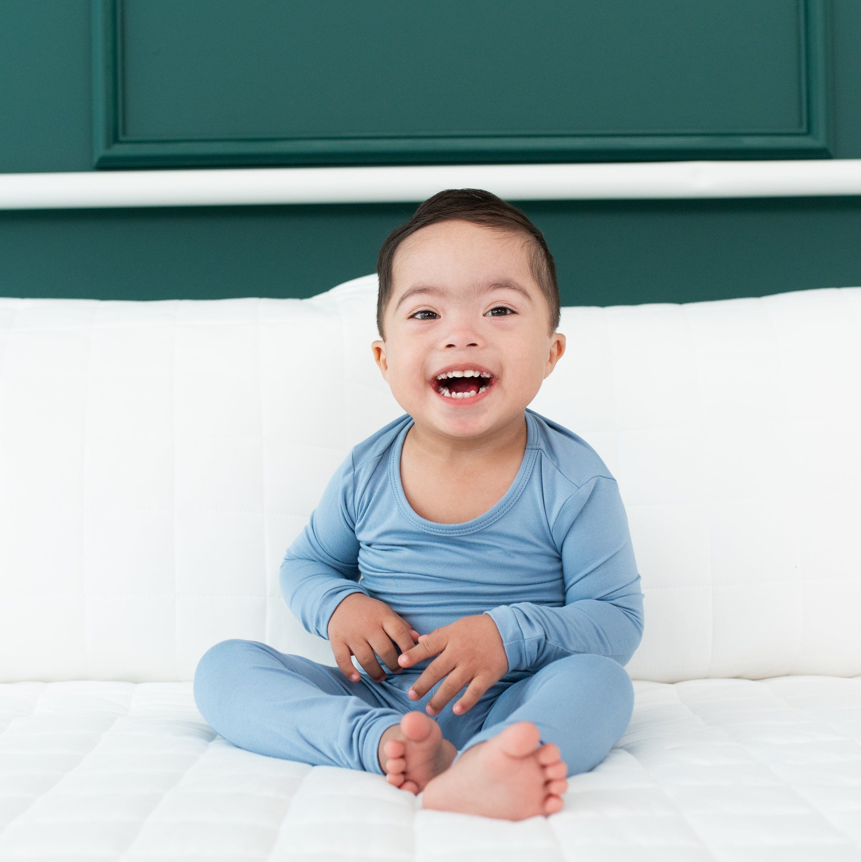 Smiling toddler sitting on a bed and wearing Long Sleeve Pajamas in Slate