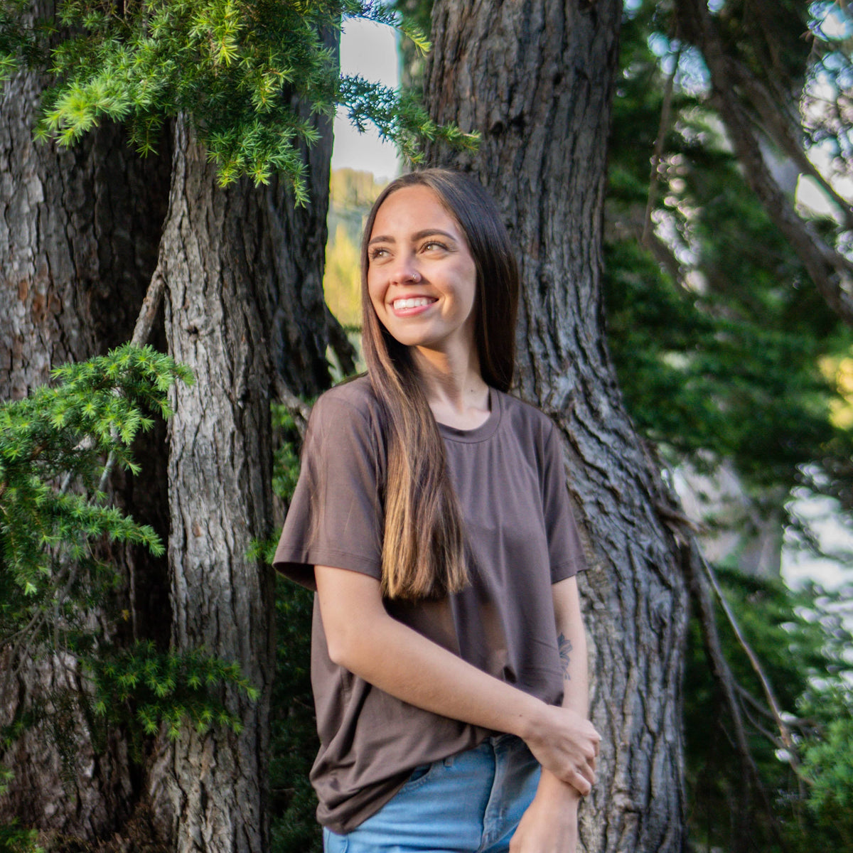 Model standing in front of trees looking off to the side wearing the Women's Crew Neck Tee in Espresso paired with jeans