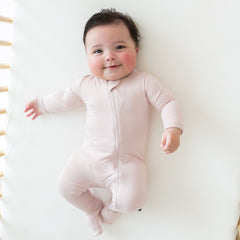 Smiling baby laying in a crib while wearing Kyte Baby Zippered Footie in Blush