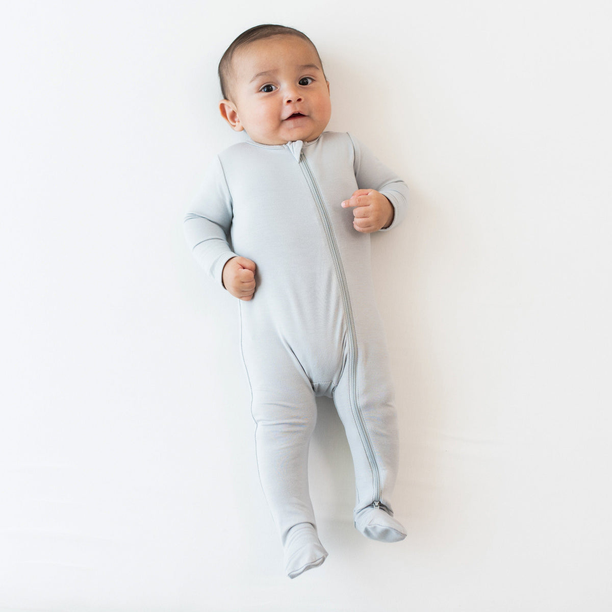 Infant laying on an off-white surface while wearing a Zippered Footie in Storm