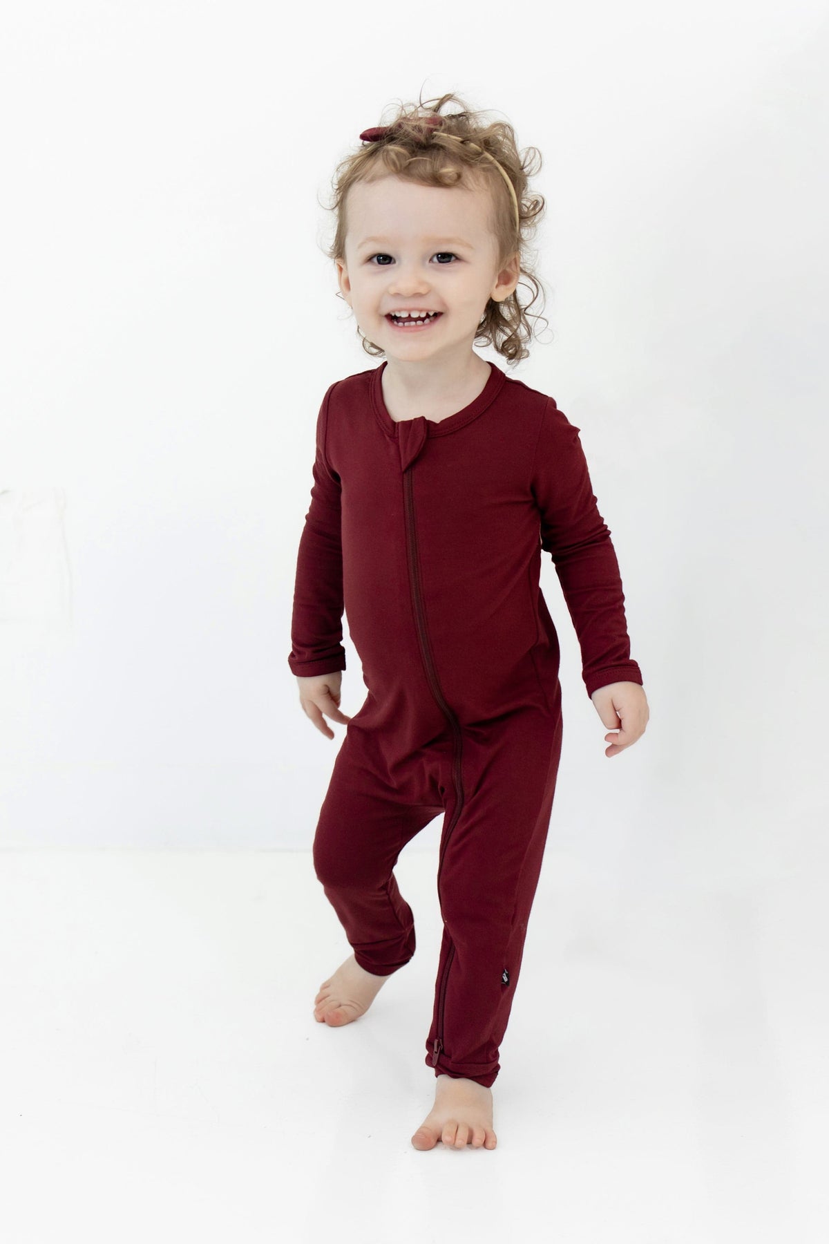 Toddler standing wearing the Zippered Romper in Burgundy with matching tiny nylon bow