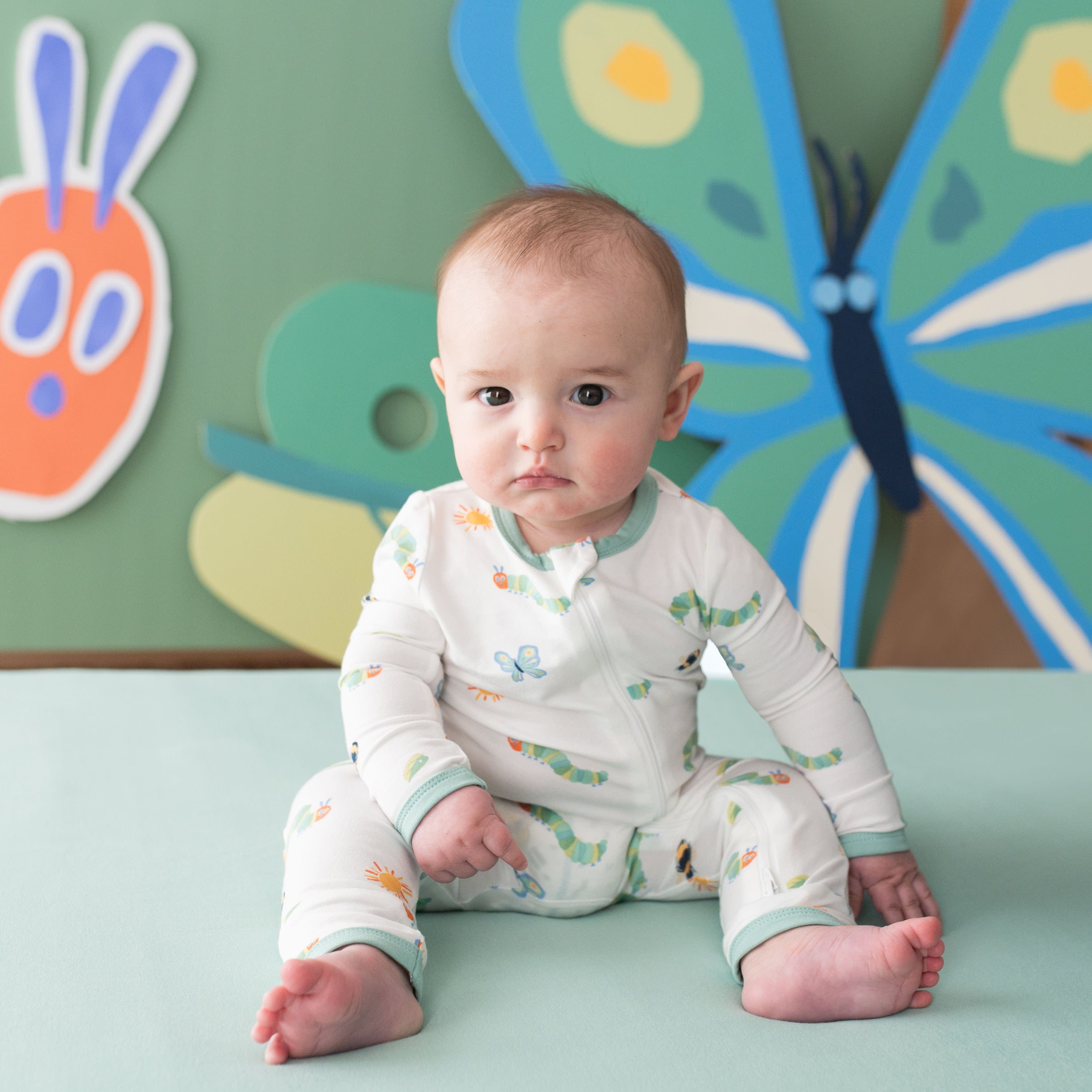 Infant siting on a green crib sheet wearing the Zippered Romper in The Very Hungry Caterpillar™ and Friends