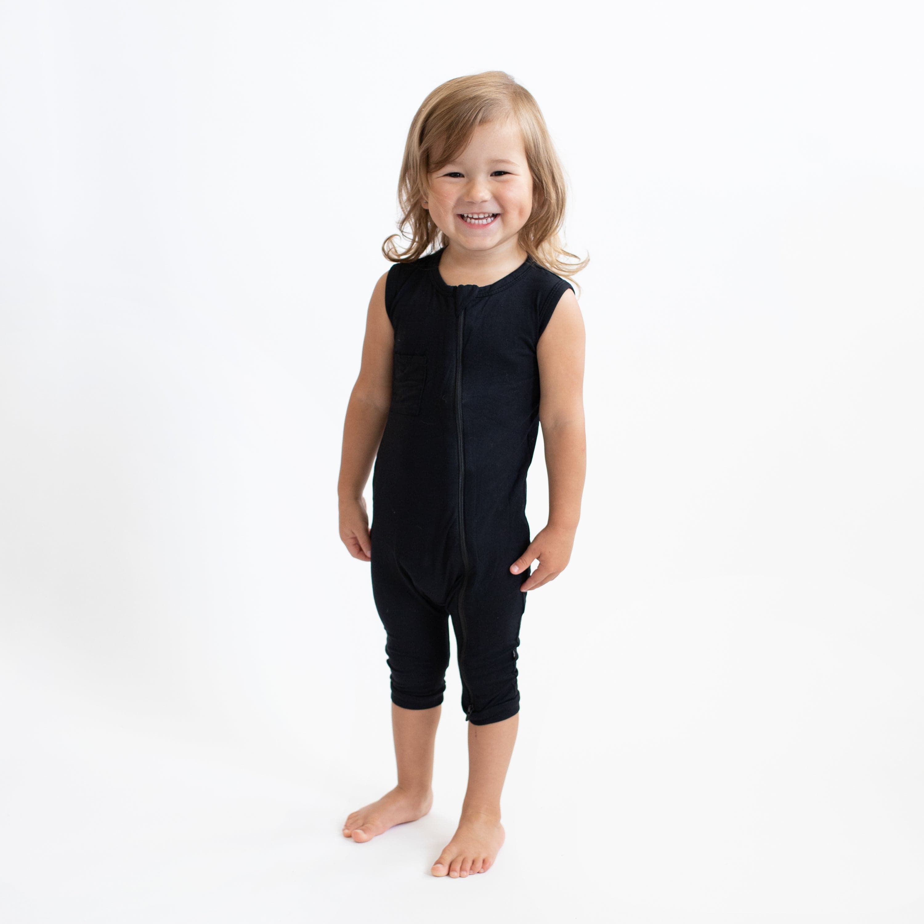 Smiling toddler standing in a white room while wearing Kyte Baby Zippered Sleeveless Romper in Midnight