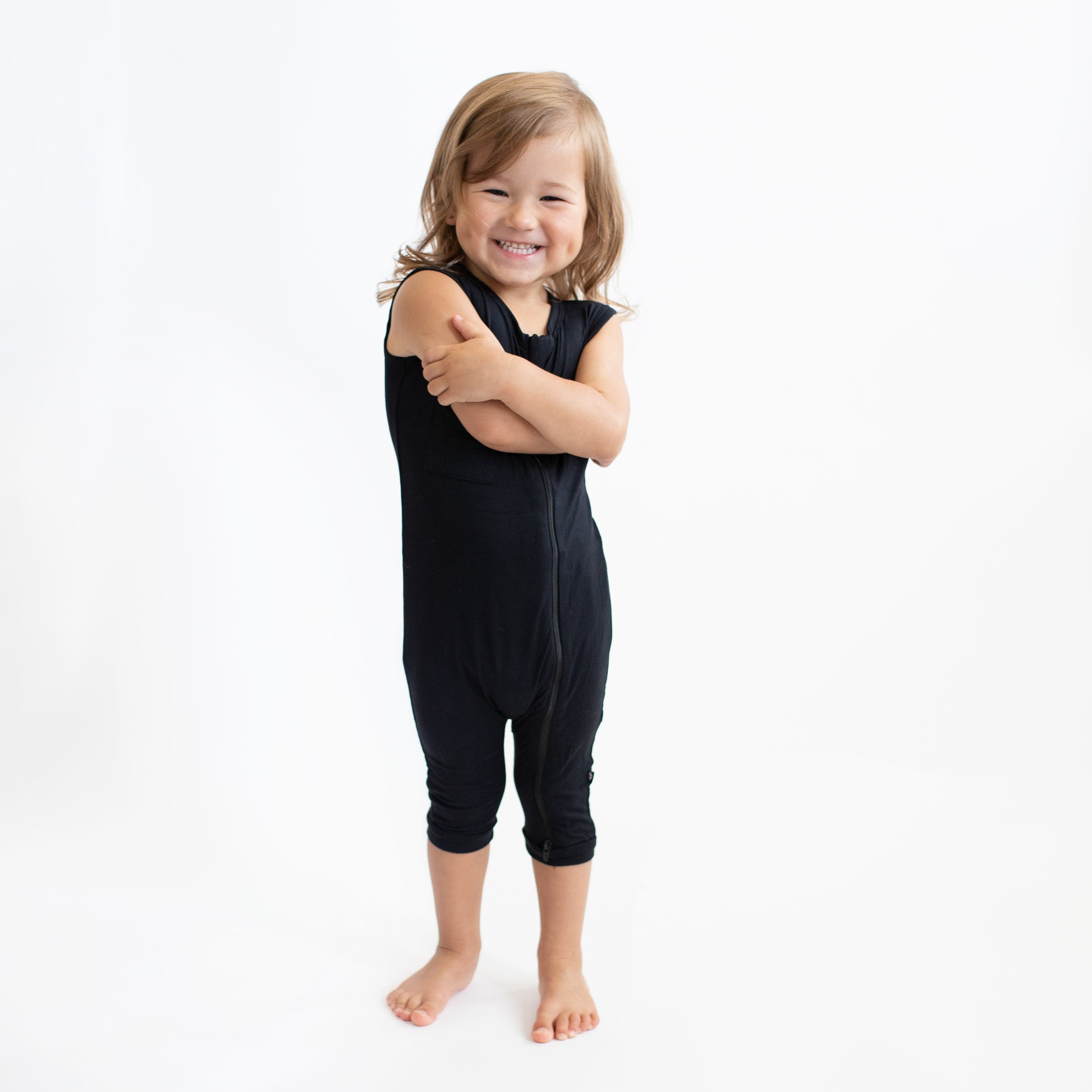 Toddler standing with arms crossed while wearing a Kyte Baby Zippered Sleeveless Romper in Midnight