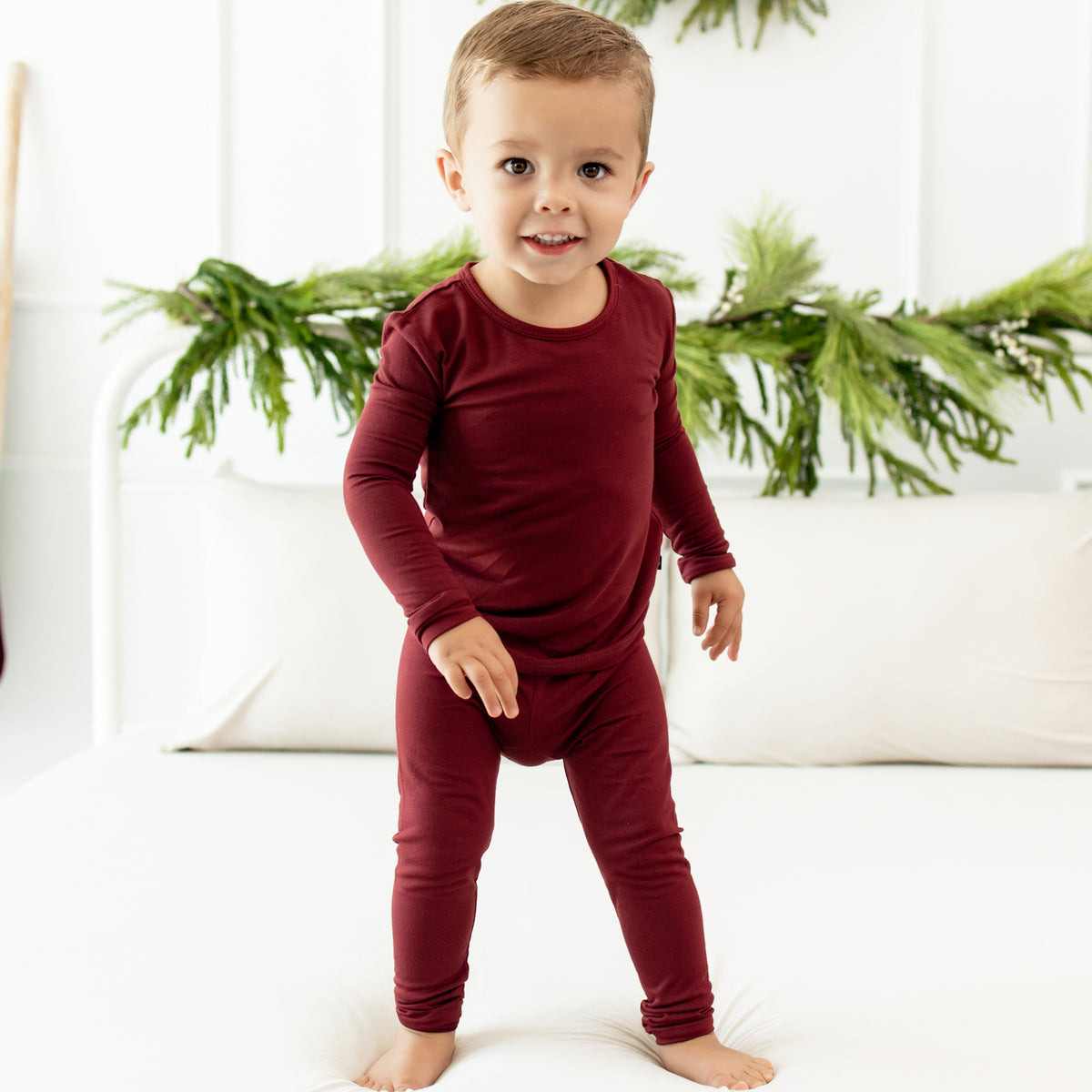 Toddler standing on a bed in front of greenery wearing the Long Sleeve Pajama Set in Burgundy