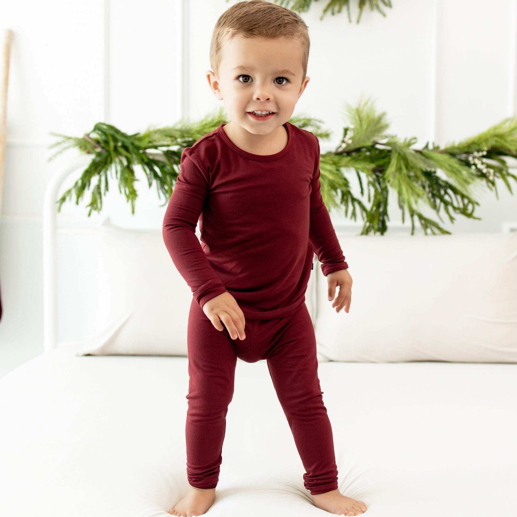 Toddler standing on a bed in front of greenery wearing the Long Sleeve Pajama Set in Burgundy