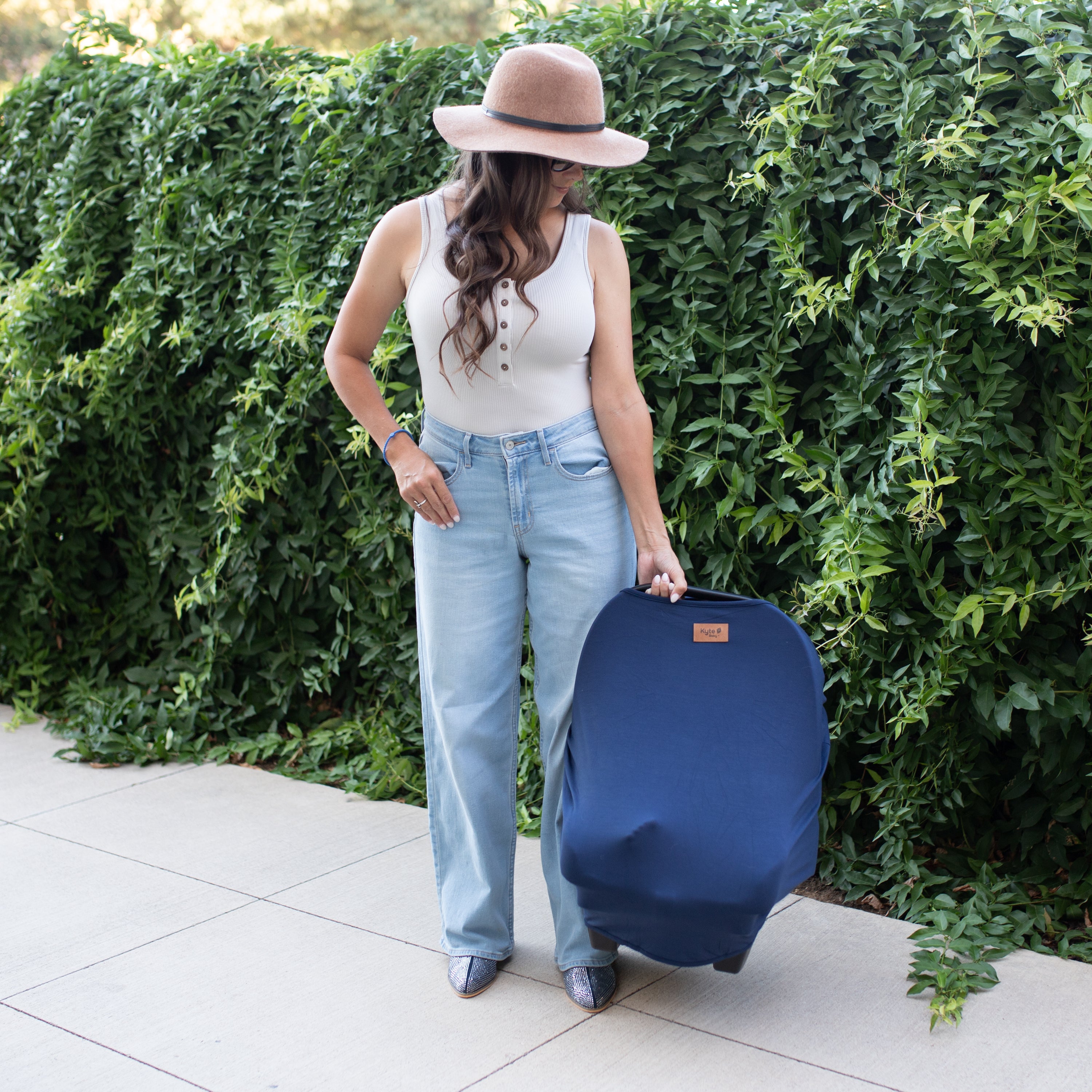 Woman holding a car seat with a navy blue over on it against a green hedge background