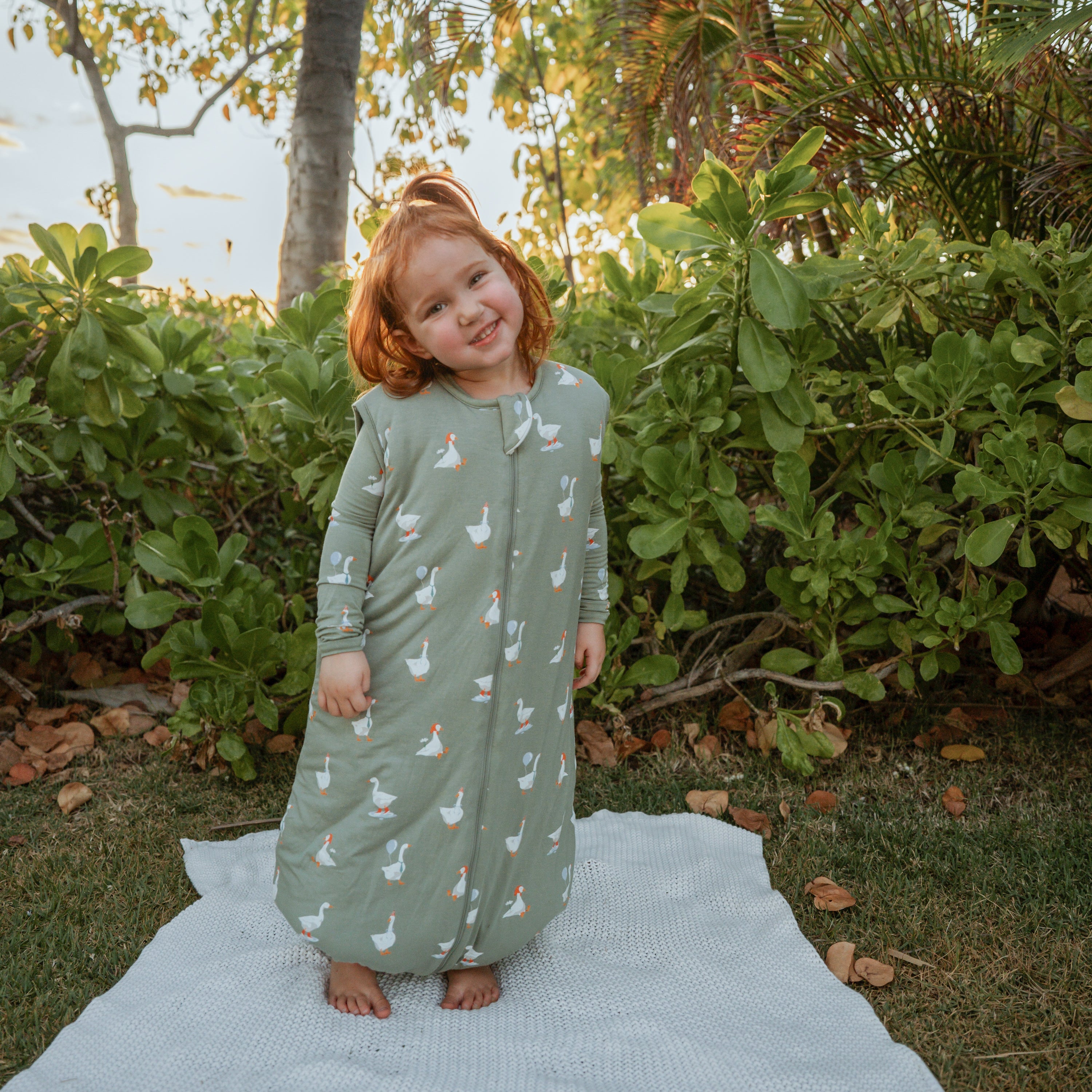 Young girl standing in a field on top of a blanket wearing the Sleep Bag Walker in Silly Goose 2.5 and matching toddler pajamas on underneath