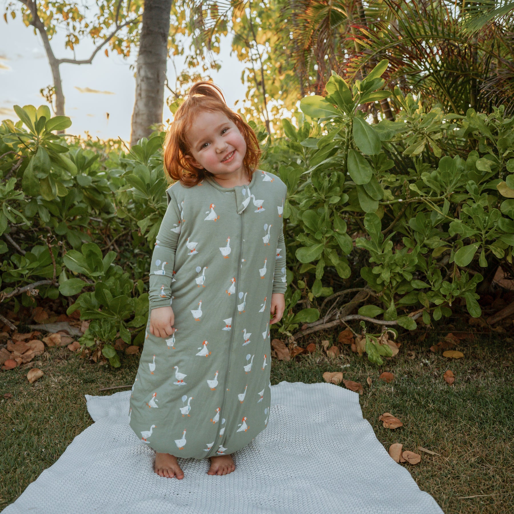 Young girl standing in a field on top of a blanket wearing the Sleep Bag Walker in Silly Goose 2.5 and matching toddler pajamas on underneath