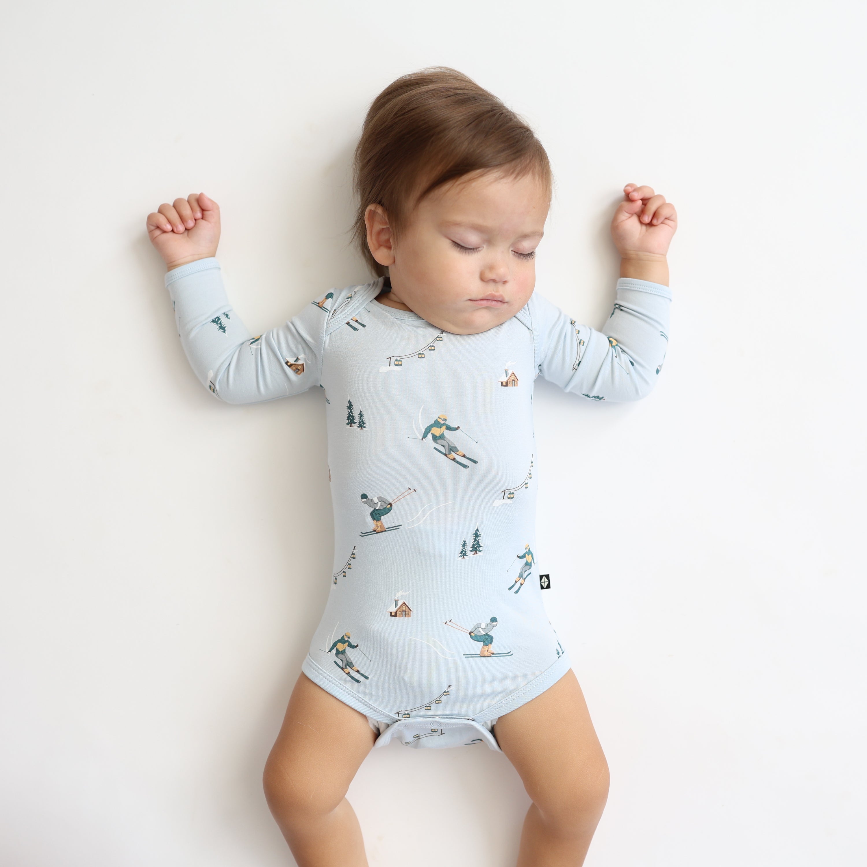 Close up of sleeping toddler laying on a light neutral surface wearing the Long Sleeve Bodysuit in Ski