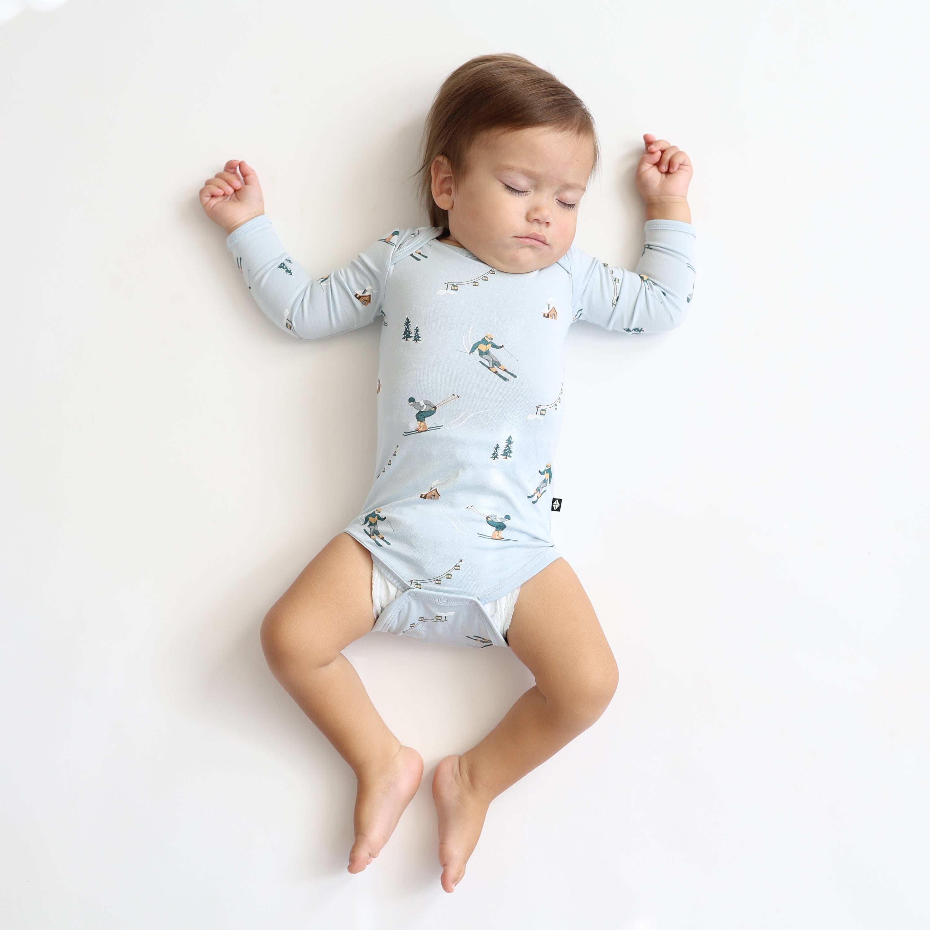 Sleeping toddler laying on a light neutral surface wearing the Long Sleeve Bodysuit in Ski