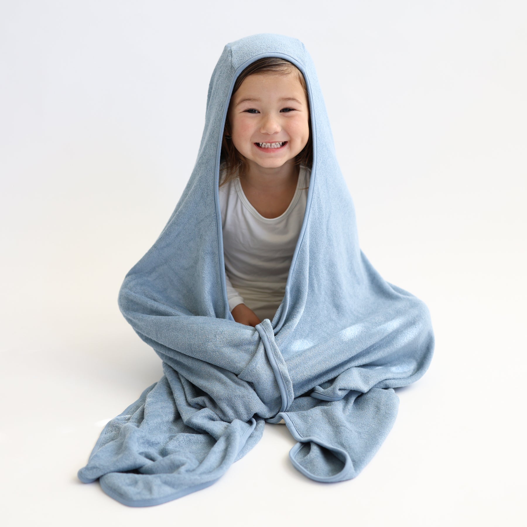 Young girl sitting on a white surface wrapped in the Toddler Hooded Bath Towel in Slate