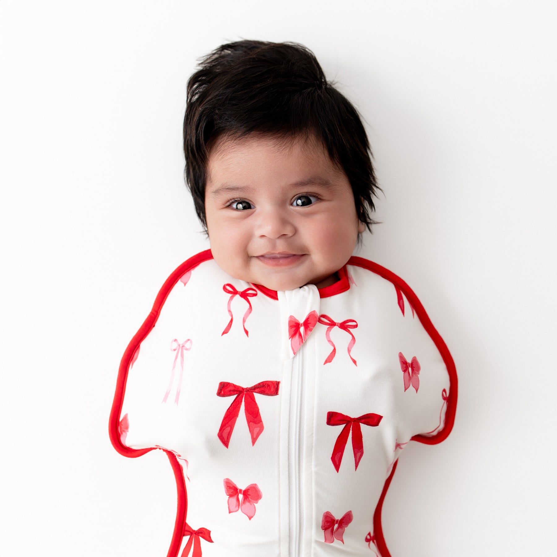 Close up of an infant laying on a white surface wearing the Swaddle Bag in Small Love Bow 0.5