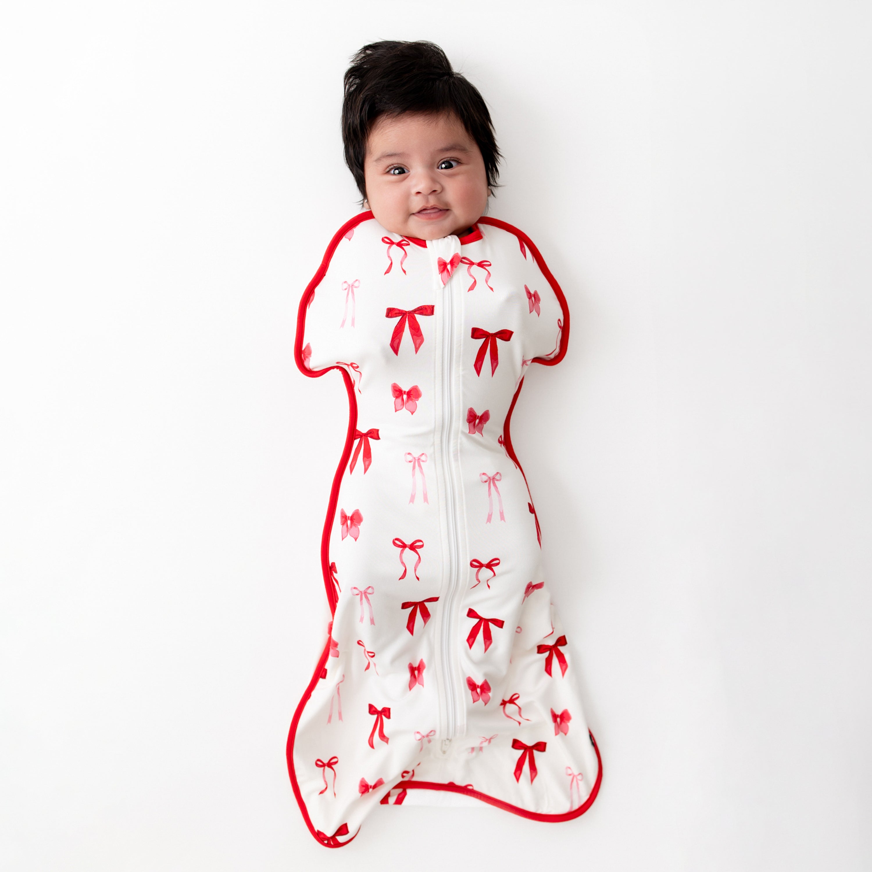 Infant laying on a white surface wearing the soft and stretchy Swaddle Bag in Small Love Bow 0.5