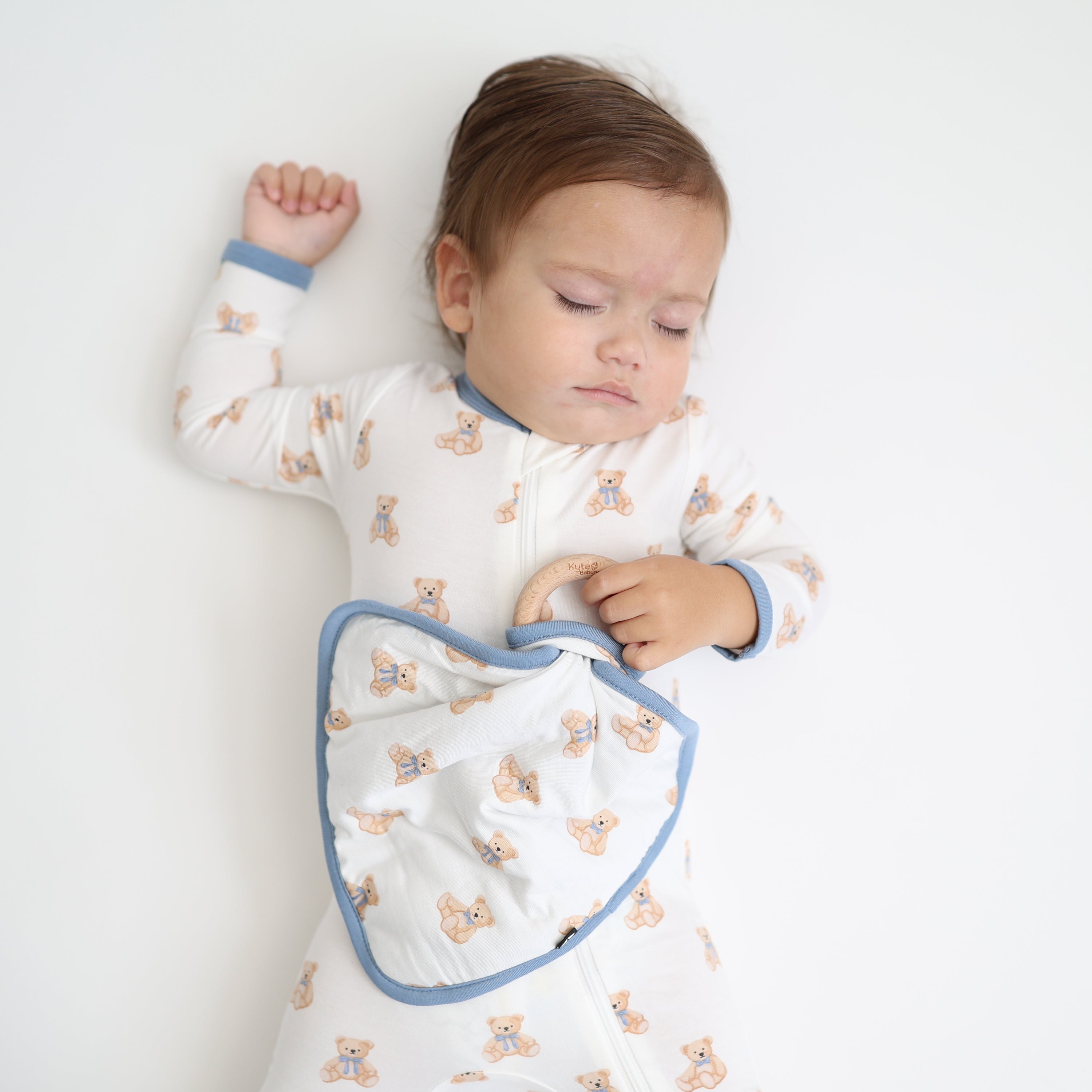 Sleeping toddler laying on a light neutral surface holding the Lovey in Teddy Bear with Removable Wooden Teething Ring wearing a matching zippered romper