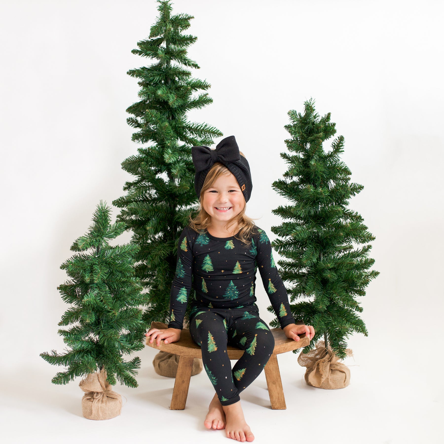 Young girl sitting on a wooden stool in front of 3 green trees wearing the Long Sleeve Pajamas in Twinkle Tree paired with a Chunky Knit Bow Headband in Black 