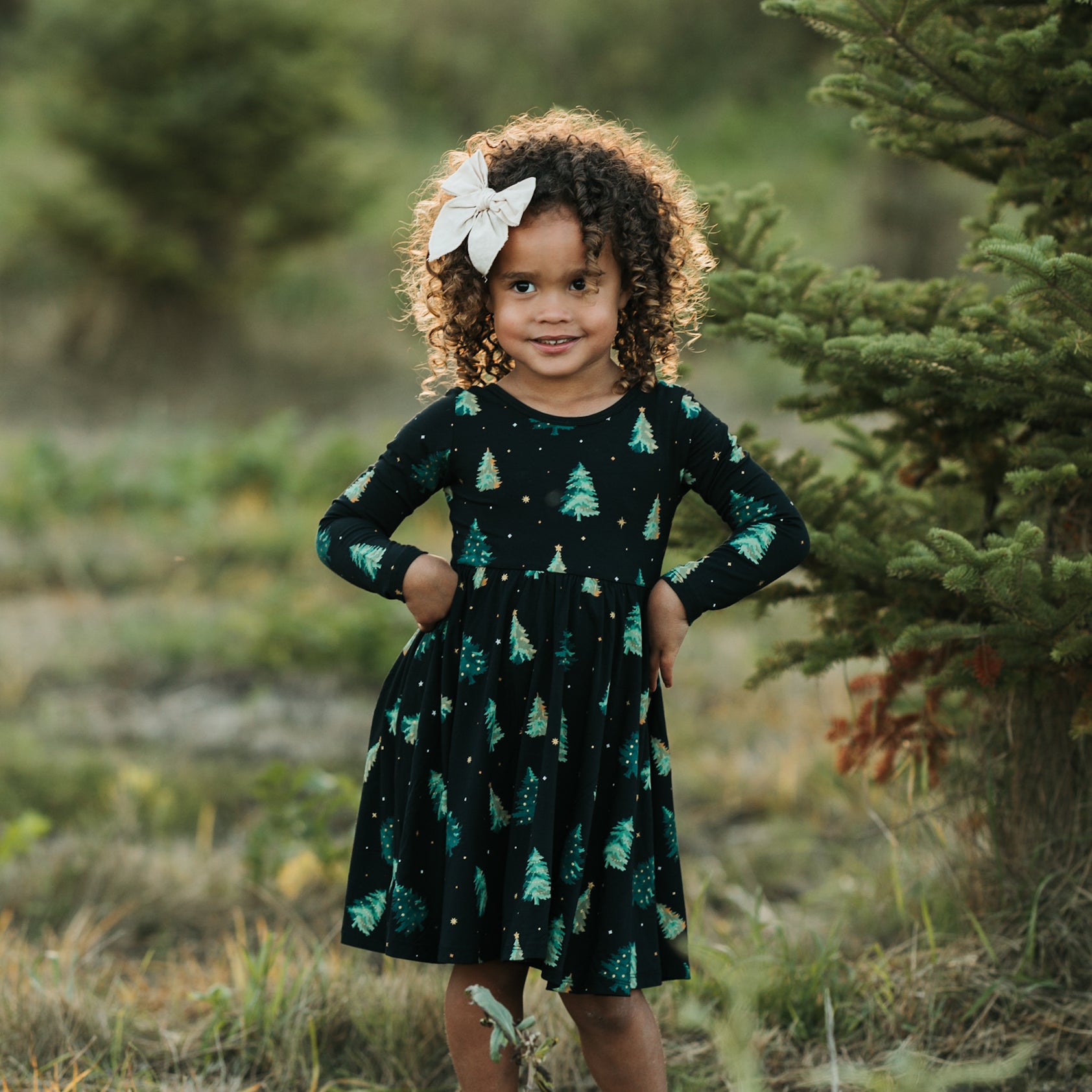 Young girl standing in a field beside a tree wearing the Long Sleeve Twirl Dress in Twinkle Tree and a white bow in her hair