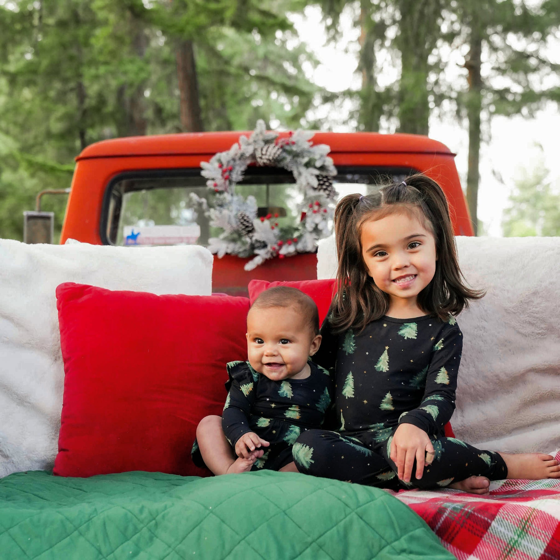 Two sisters sitting in the back of a truck bed with white, red and green pillows and blankets wearing the Long Sleeve Pajamas in Twinkle Tree and matching long sleeve twirl bodysuit dress