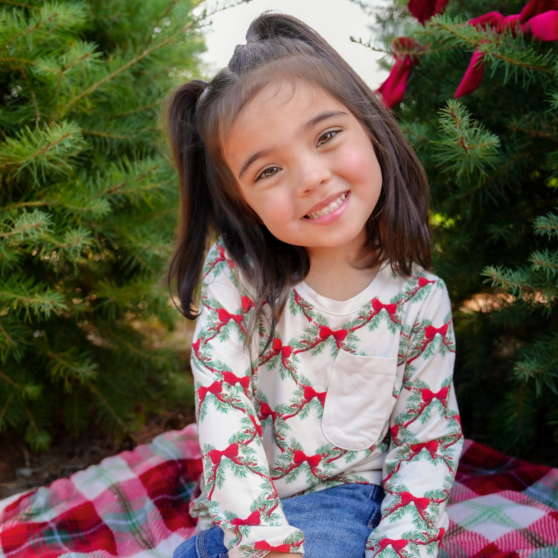 Young girl sitting on a red plaid blanket between two trees wearing the Long Sleeve Toddler Crew Neck Tee in Holiday Bow