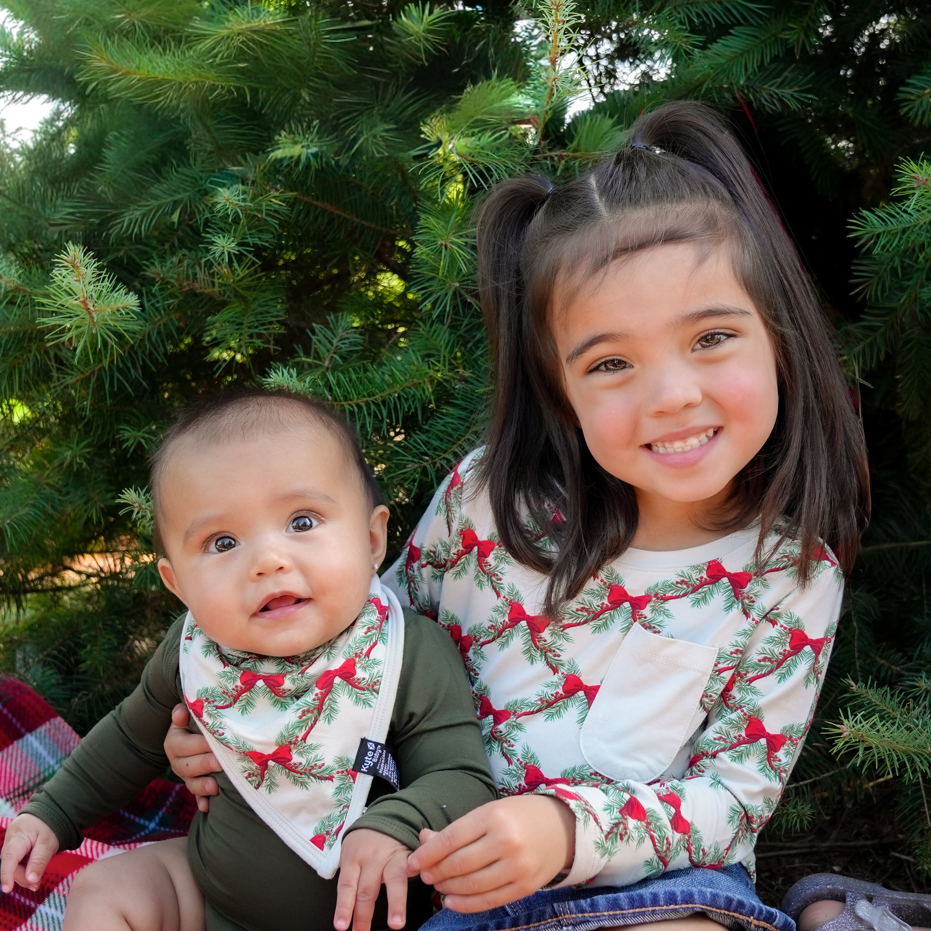 Two sisters sitting side by side in front of a tree matching in Holiday Bow. Older sister is wearing the Holiday Bow Long Sleeve Crew Neck tee and youngest is wearing the Bib in Holiday Bow and a Fir Long Sleeve Bodysuit