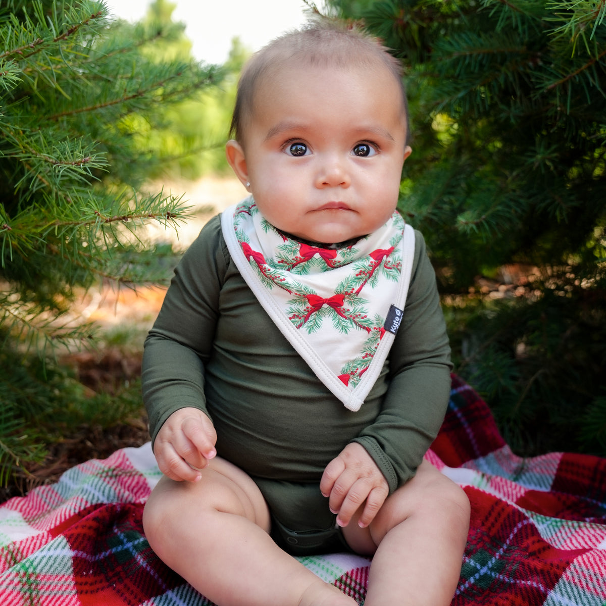Infant sitting on a plaid blanket between two trees wearing the Bib in Holiday Bow and a dark green long sleeve bodysuit
