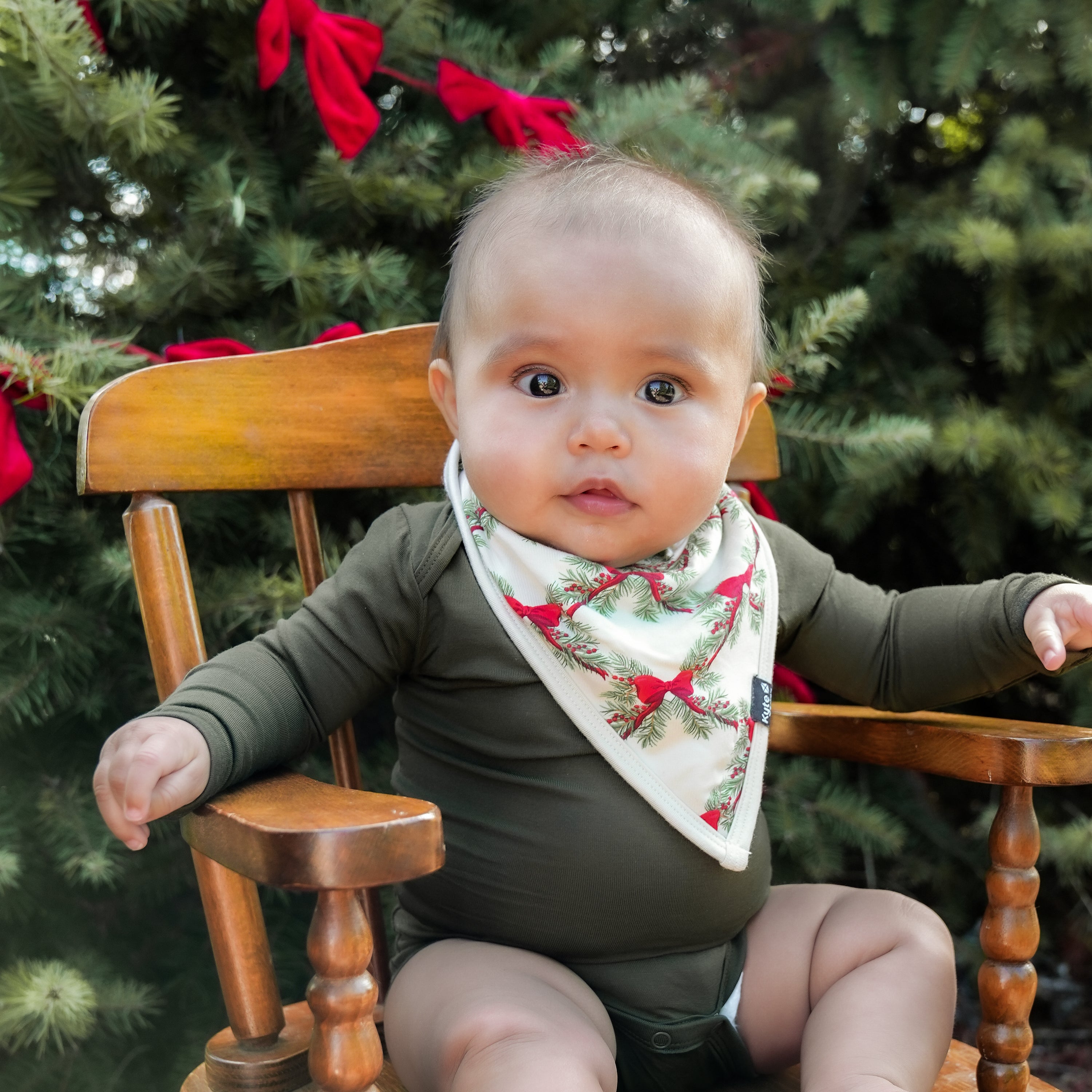 Infant sitting in a wooden chair in front of trees wearing the Bib in Holiday Bow and Fir long Sleeve Bodysuit